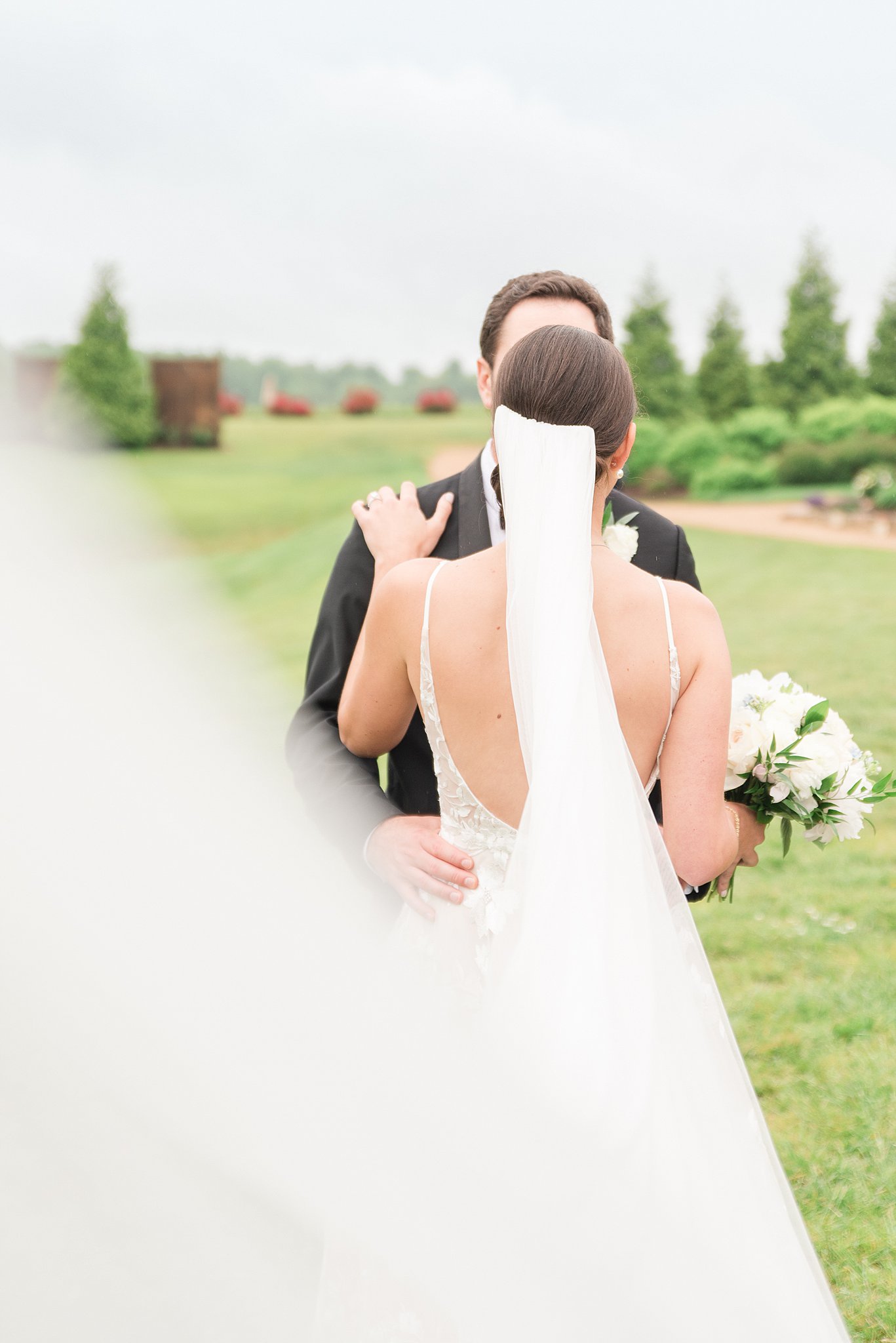 Newlyweds stand in an open lawn holding each other while the vail flies behind the bride