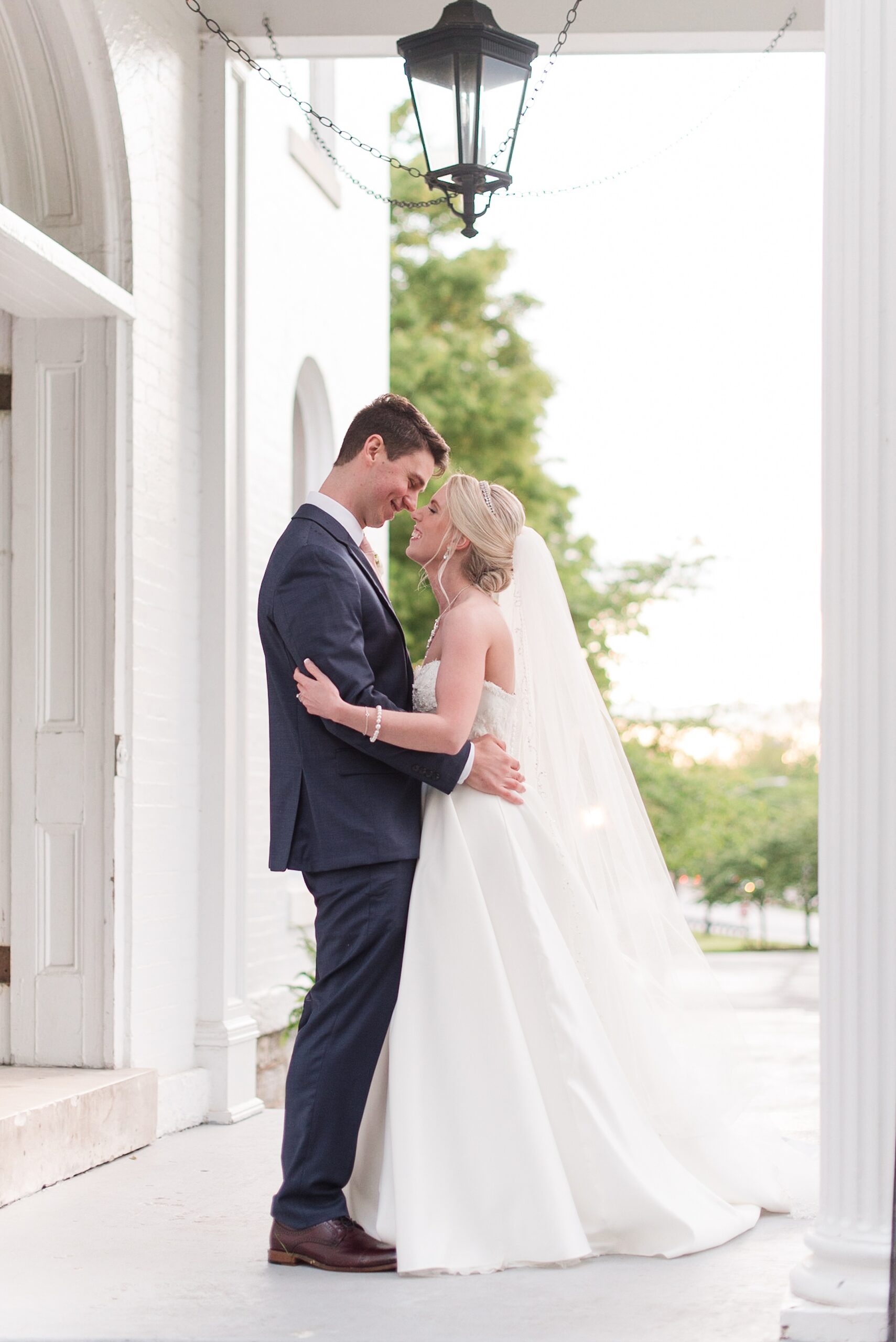 Newlyweds nuzzle noses while standing on a porch at sunset