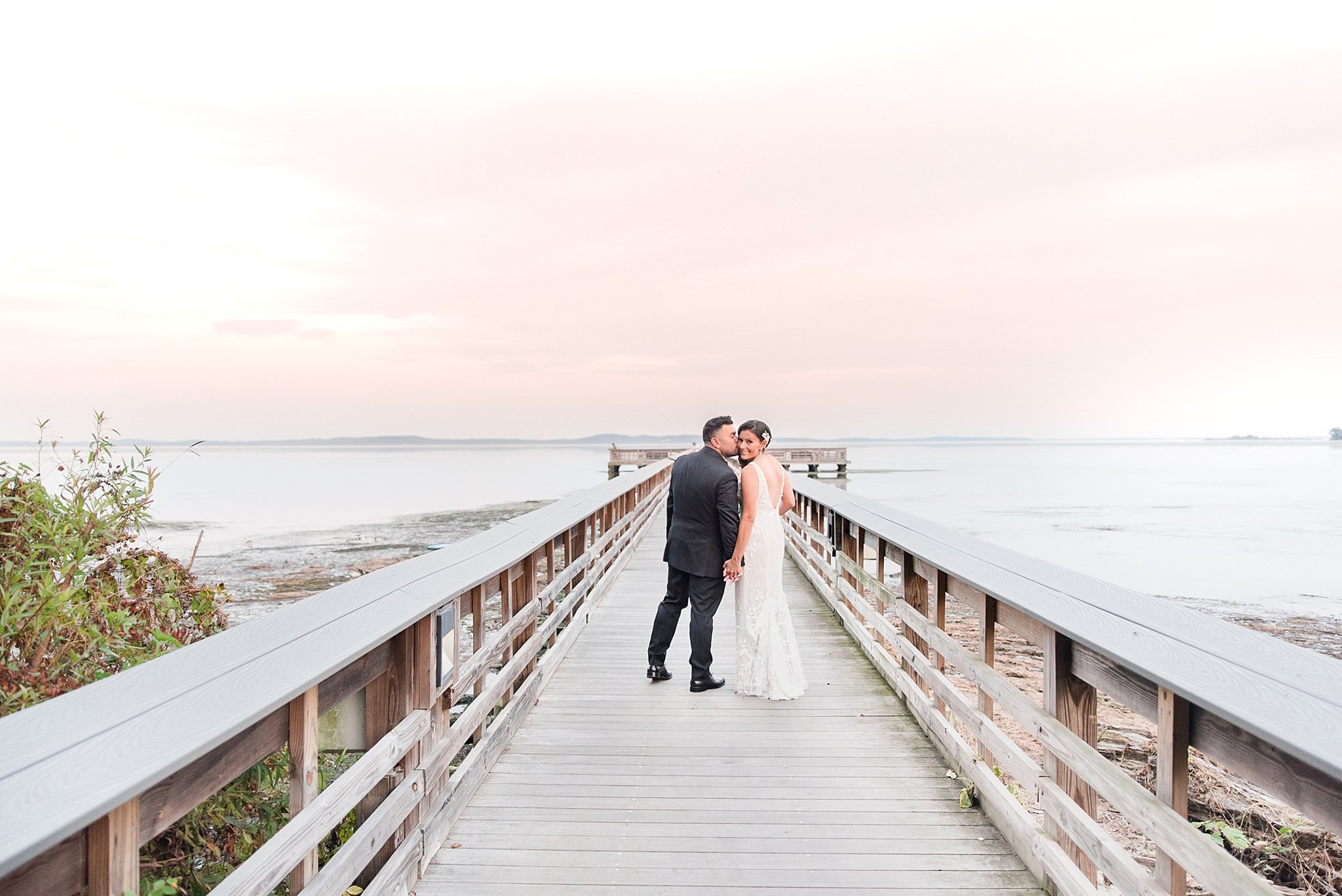 A groom kisses his bride as they walk up a long wooden boardwalk