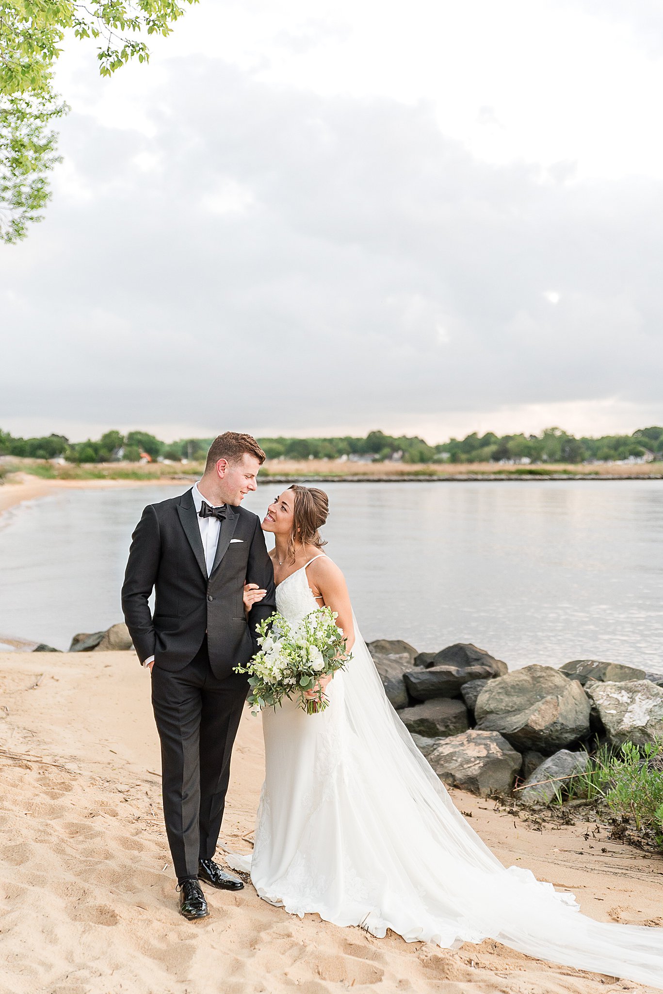A bride hangs on the arm of her groom while walking on a sandy beach at one of the incredible Baltimore Waterfront Wedding Venues