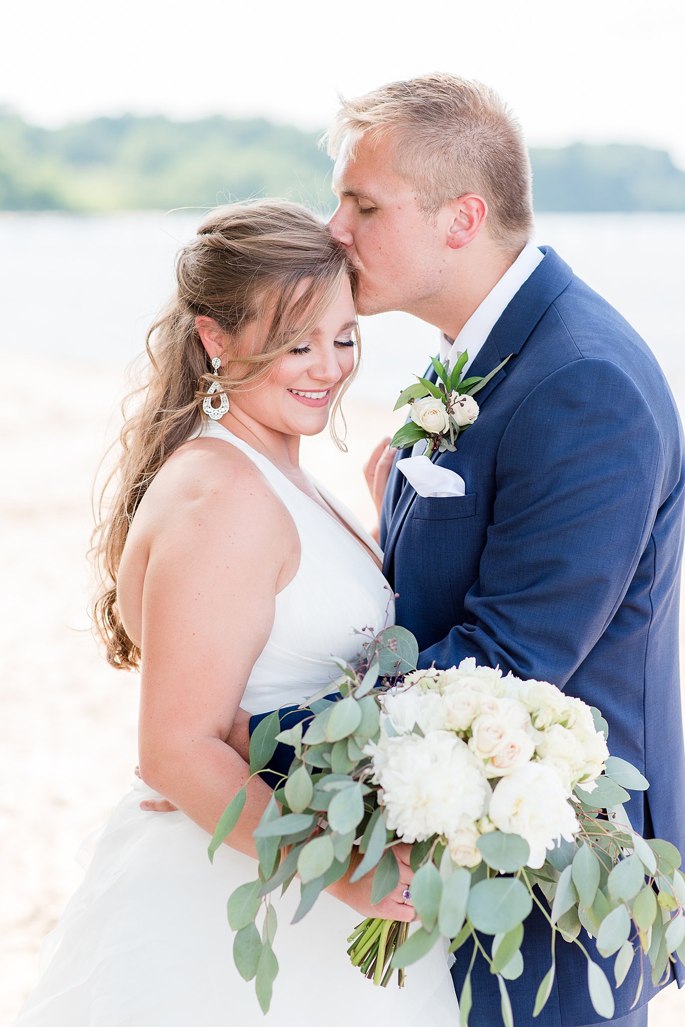 A groom kisses his bride while wearing a blue suit by a large lake at one of the Baltimore Waterfront Wedding Venues