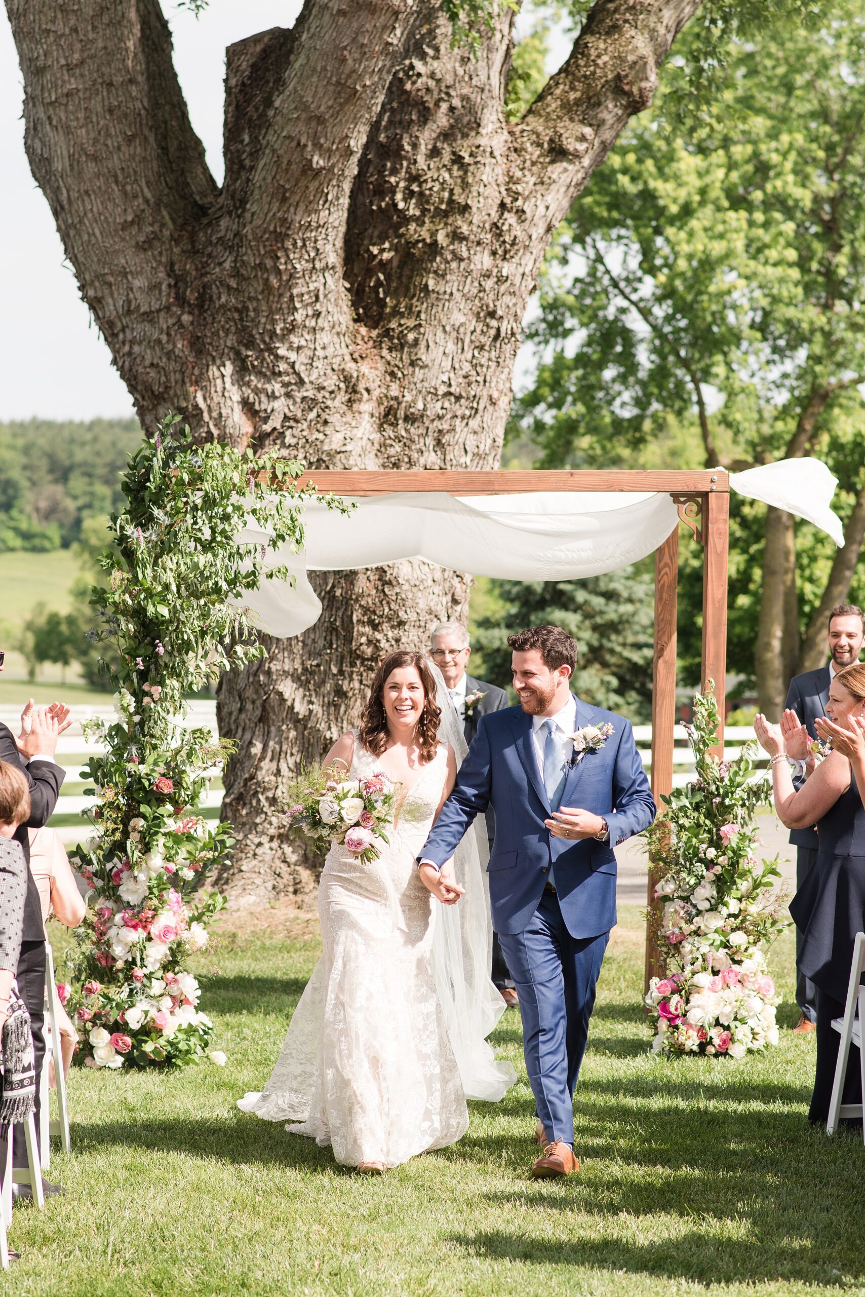 Newlyweds smile while holding hands and exiting their outdoor wedding ceremony to applause