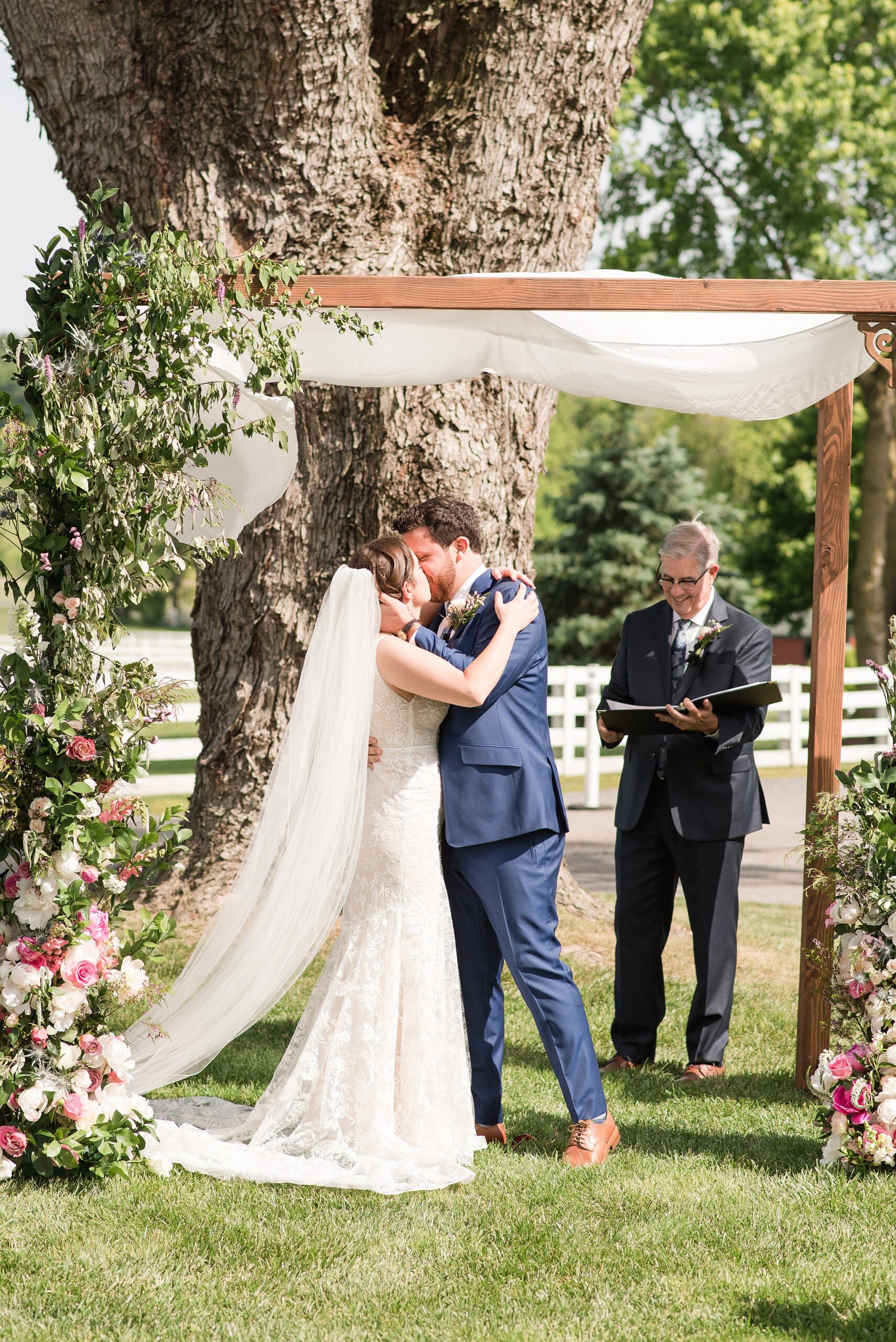 Newlyweds kiss to end their Bluebird Manor wedding ceremony under a wooden arbor covered in flowers