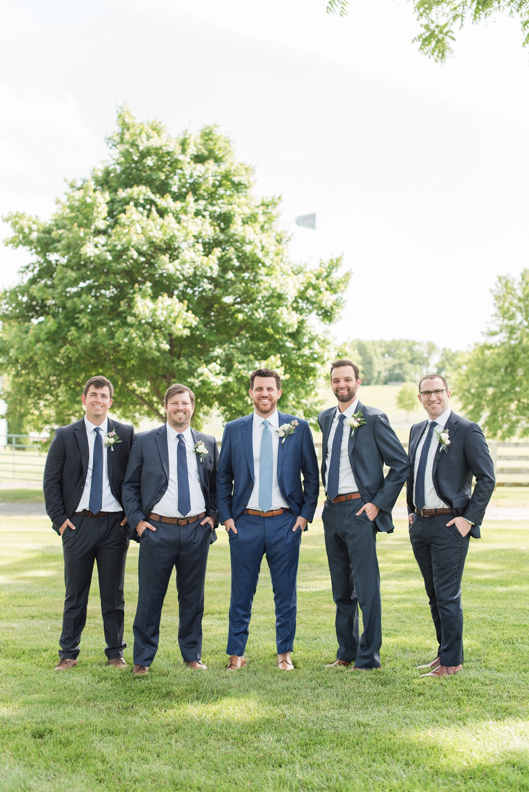 A groom in a blue suit stands with his groomsmen in a field in grey suits