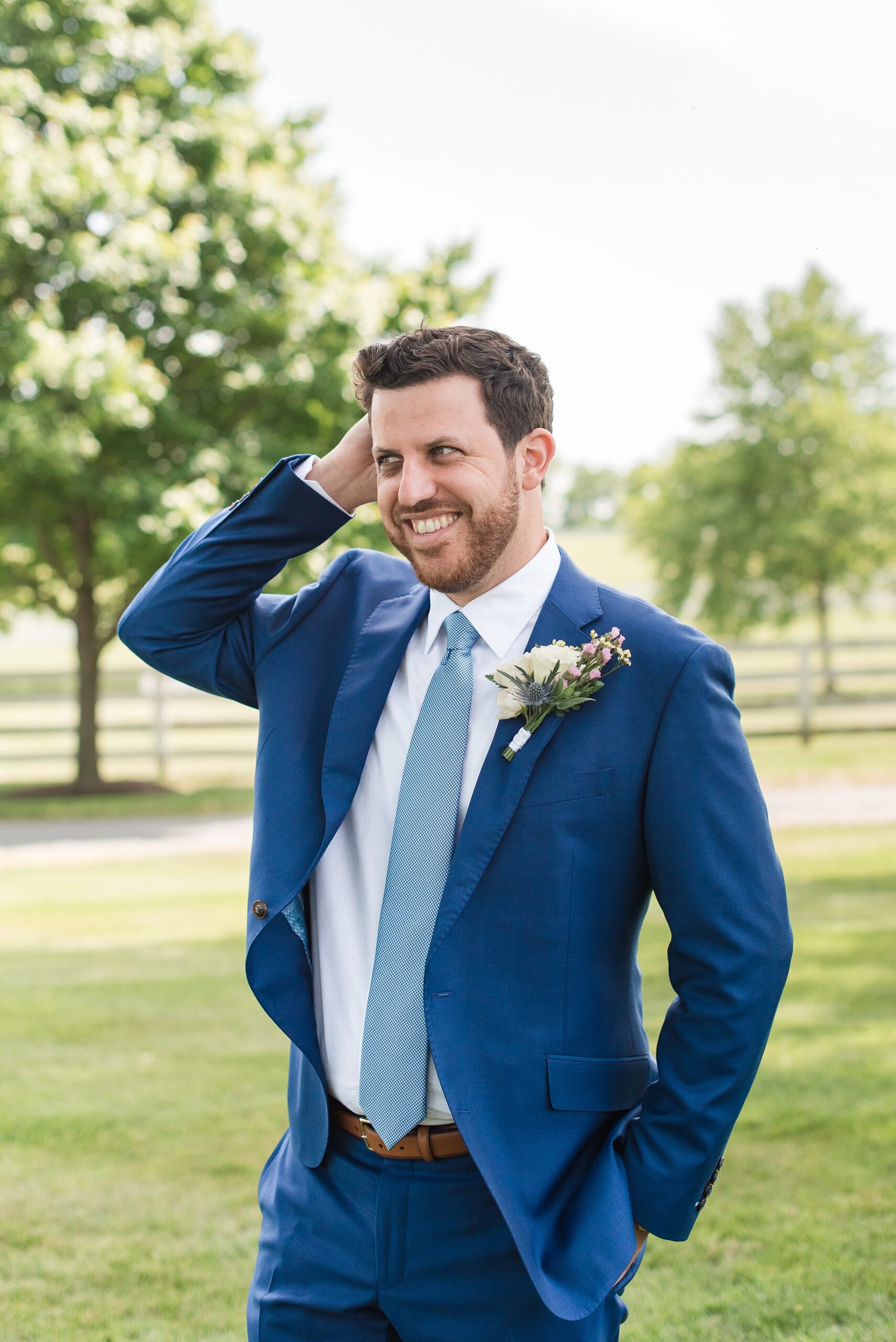 A groom in a blue suit runs a hand through his hair while standing in a lawn