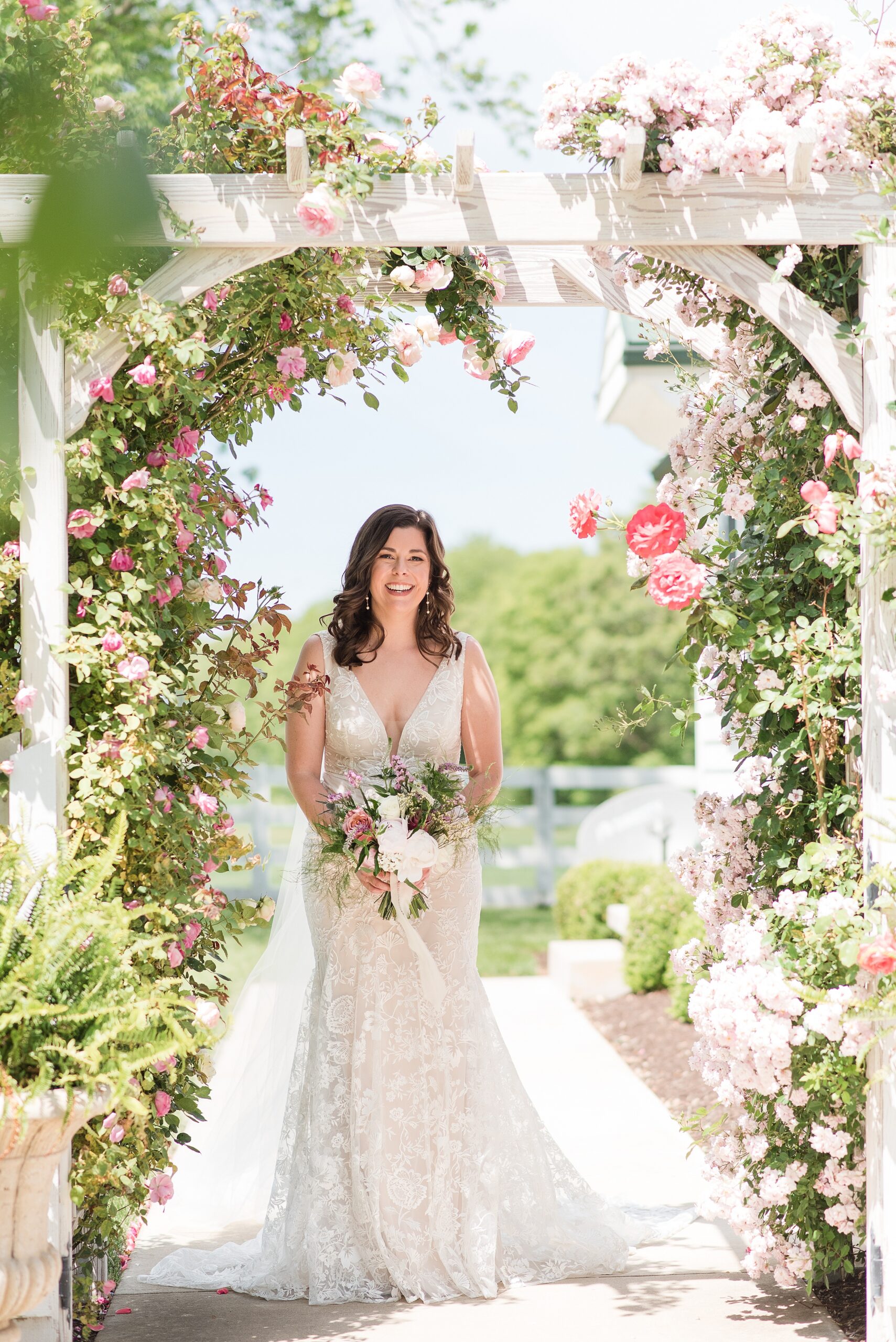 A bride in a lace dress stands under a flower covered garden arch at the Bluebird Manor