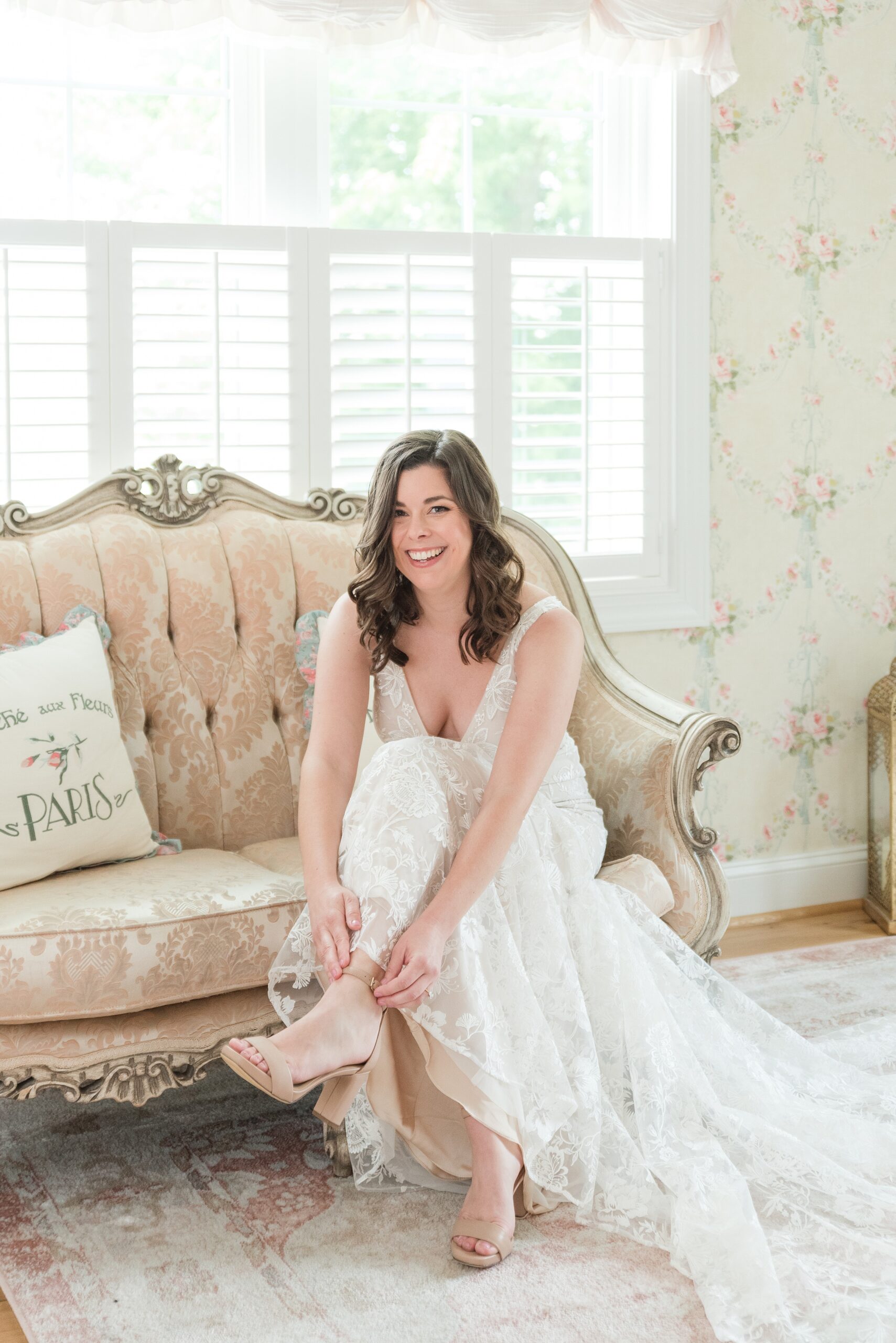 A bride puts on her shoes while sitting on a vintage couch under a window