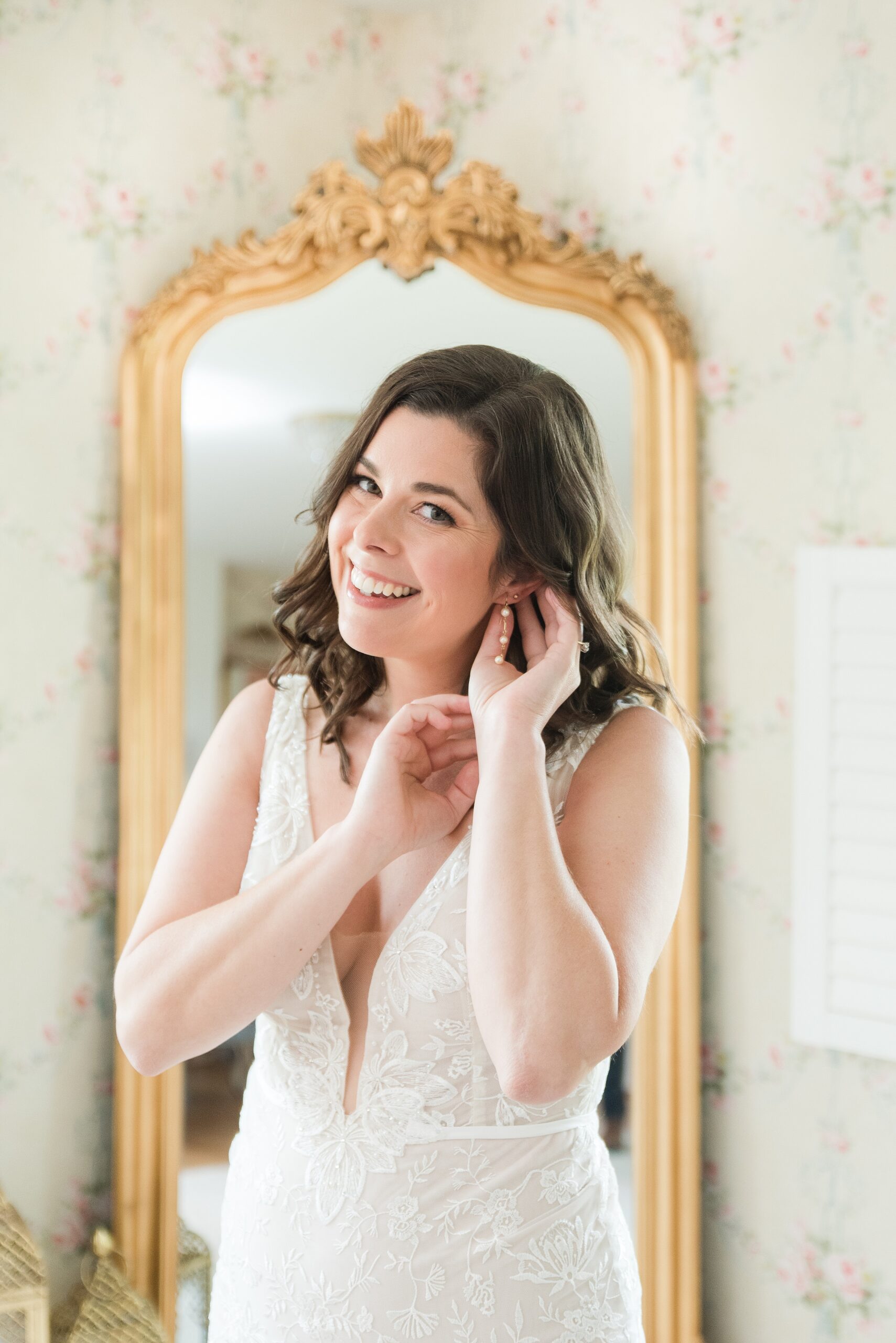 A bride in a lace dress puts on her earrings in front of a mirror at her Bluebird Manor wedding