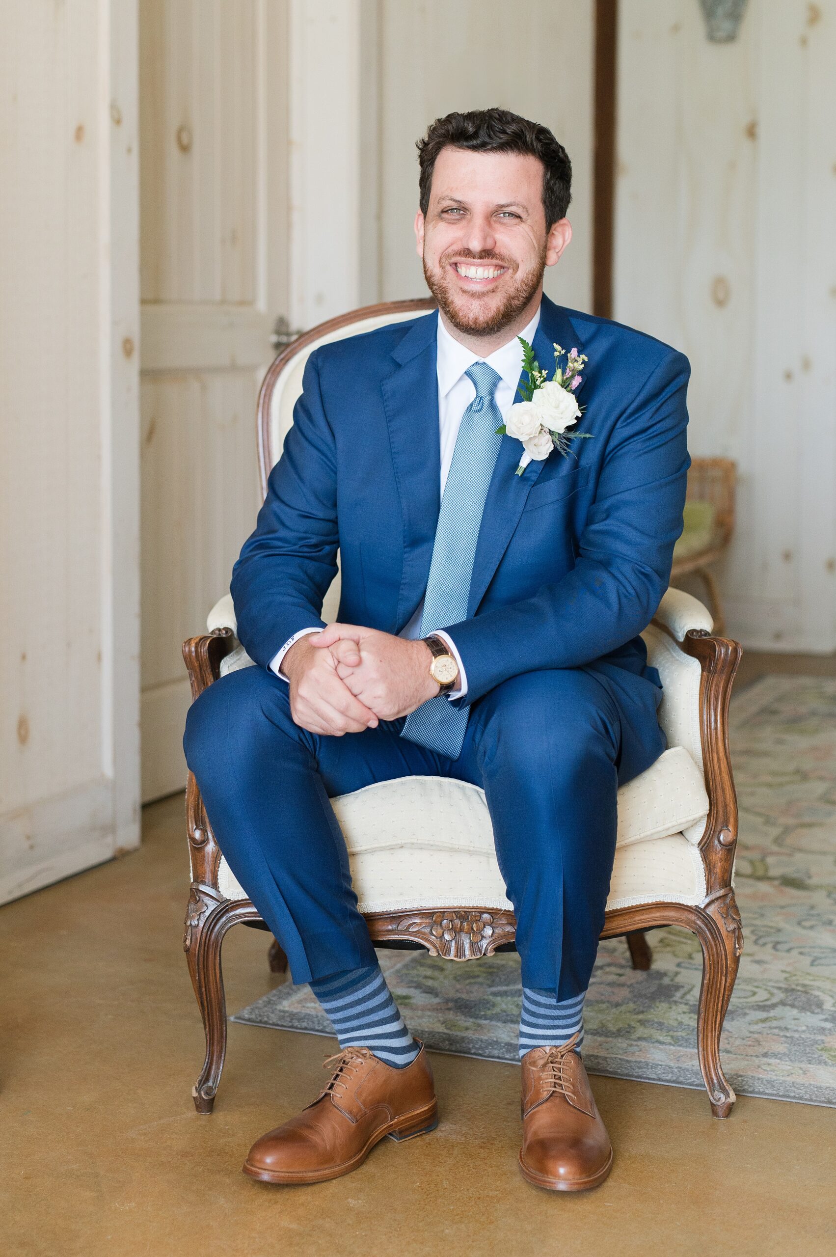 A groom sits in an antique chair smiling big in a blue suit