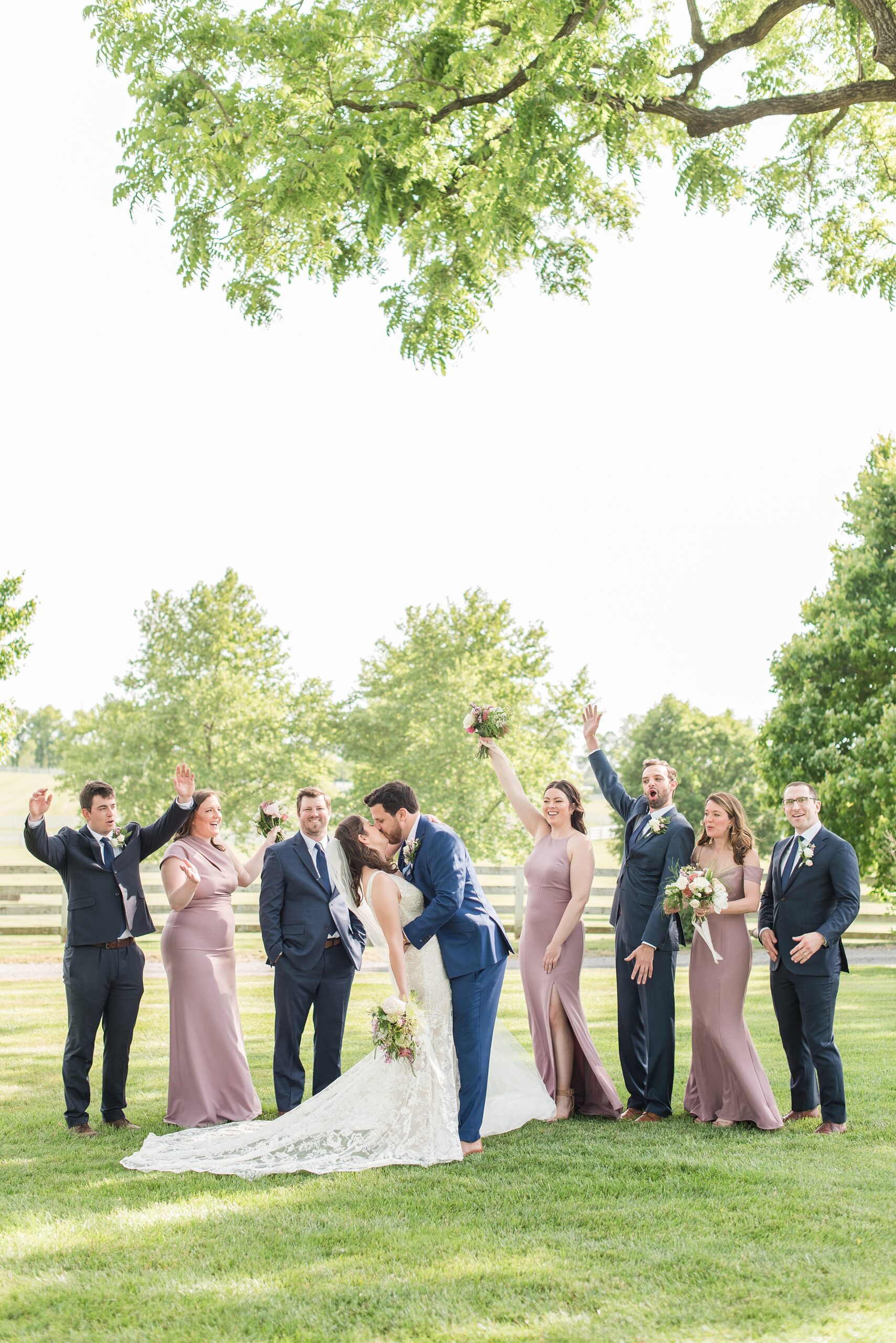 Newlyweds kiss while celebrating in a grassy lawn with their wedding party celebrating