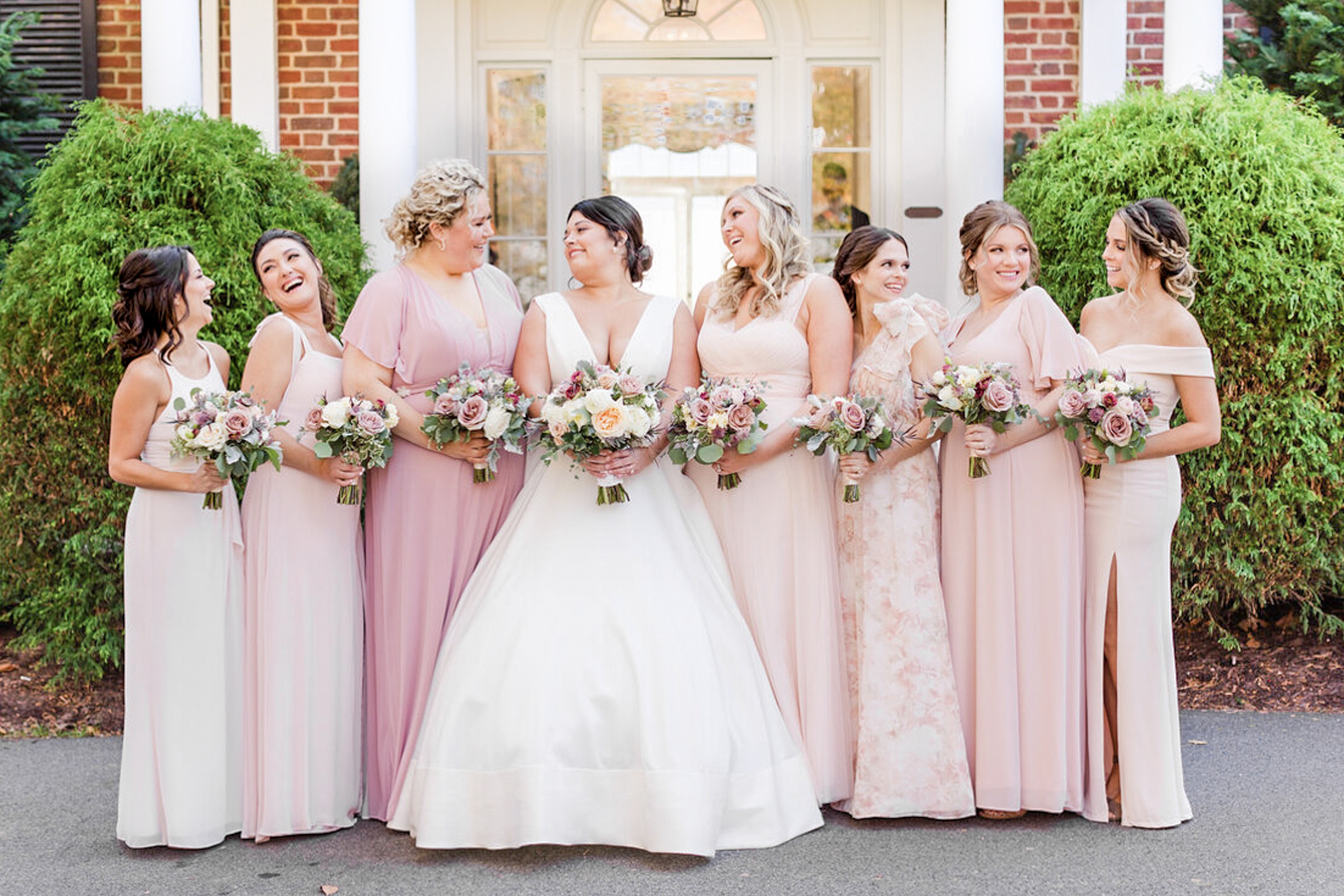 A bride stands at the front door of a brick building laughing with her bridesmaids