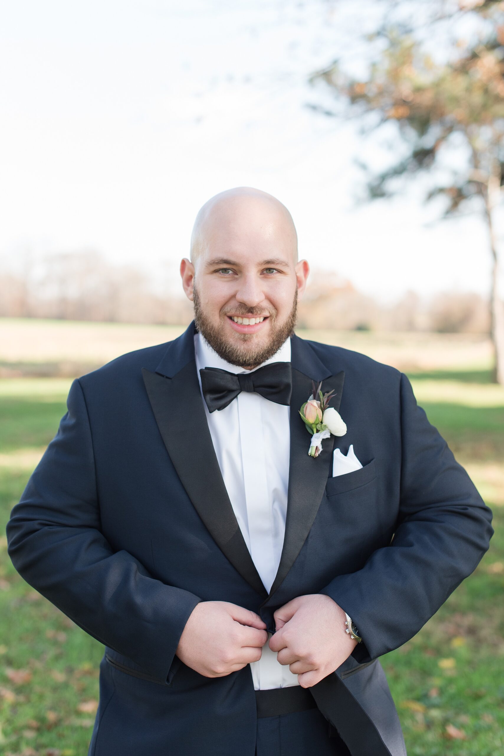 A groom smiles while buttoning his black tuxedo jacket in a grassy field