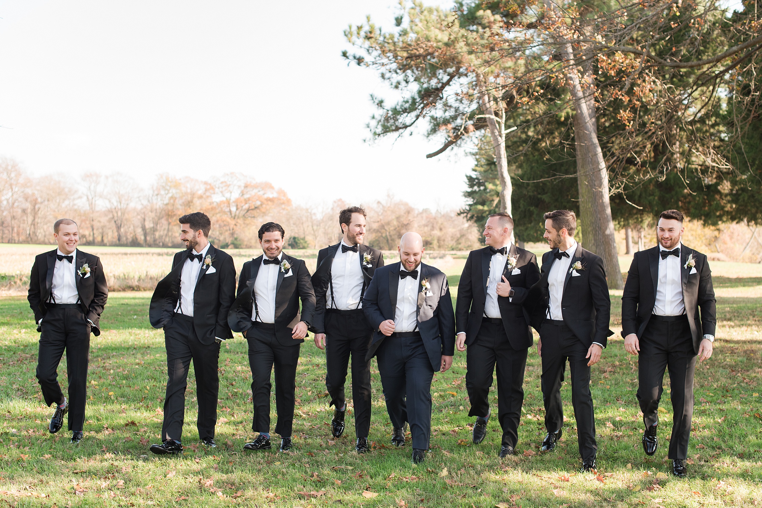 A groom in a black tux walks through a lawn with his many groomsmen in matching tuxes