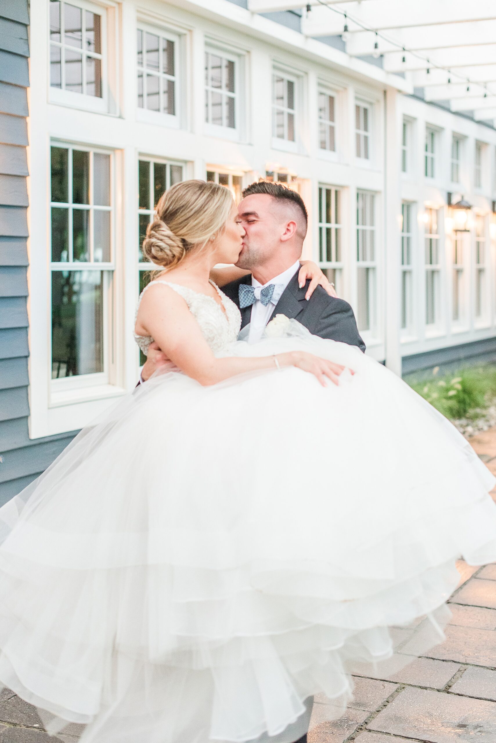 A groom carries and kisses his bride while walking on a patio under a pergola at their Chesapeake Bay Beach Club Wedding