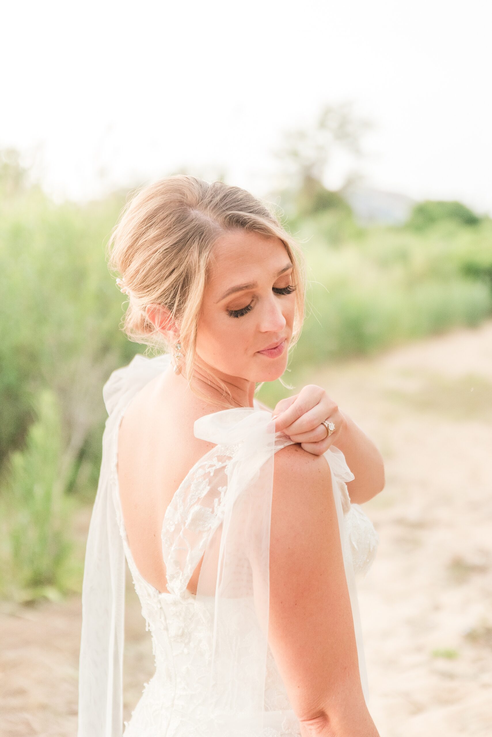 A bride adjusts her shoulder bow while walking a path at sunset