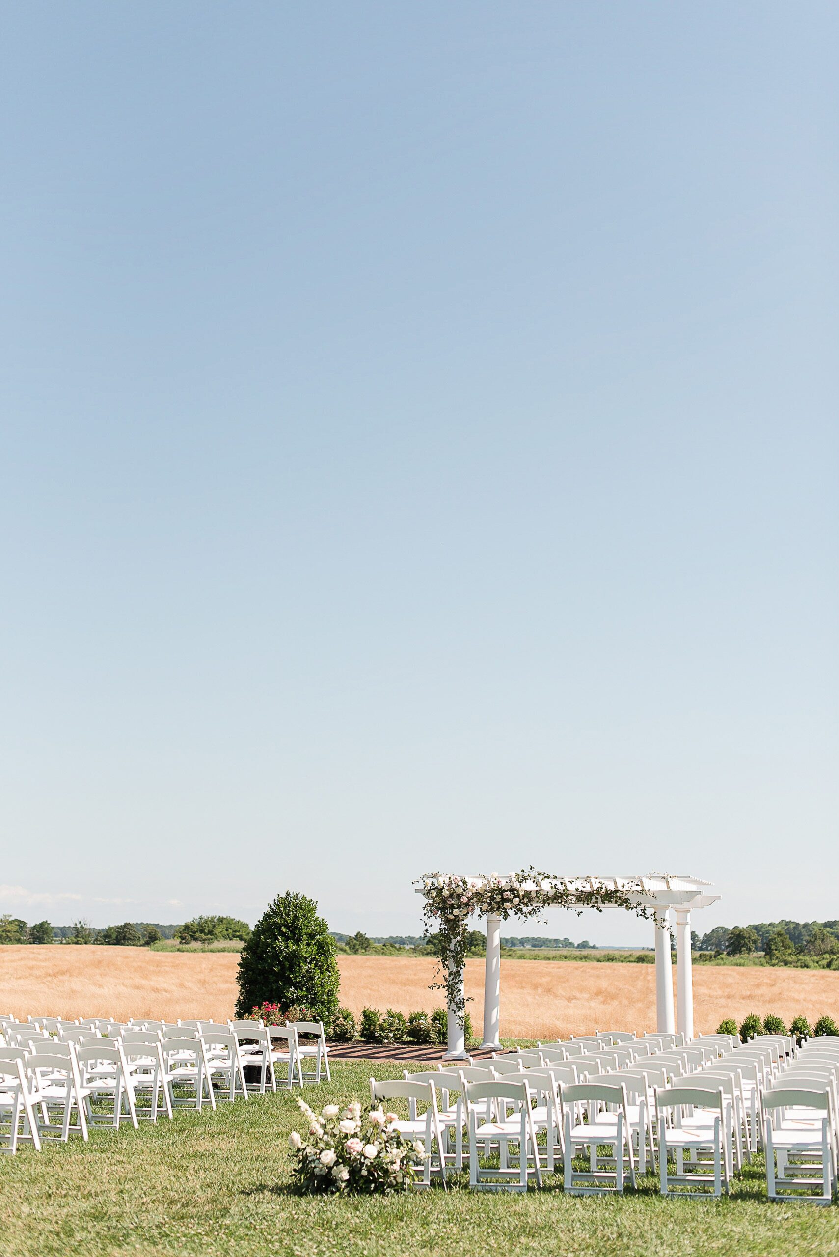 A view of a Kent Island Resort Wedding ceremony set up with white column pergola and white chairs in an open field
