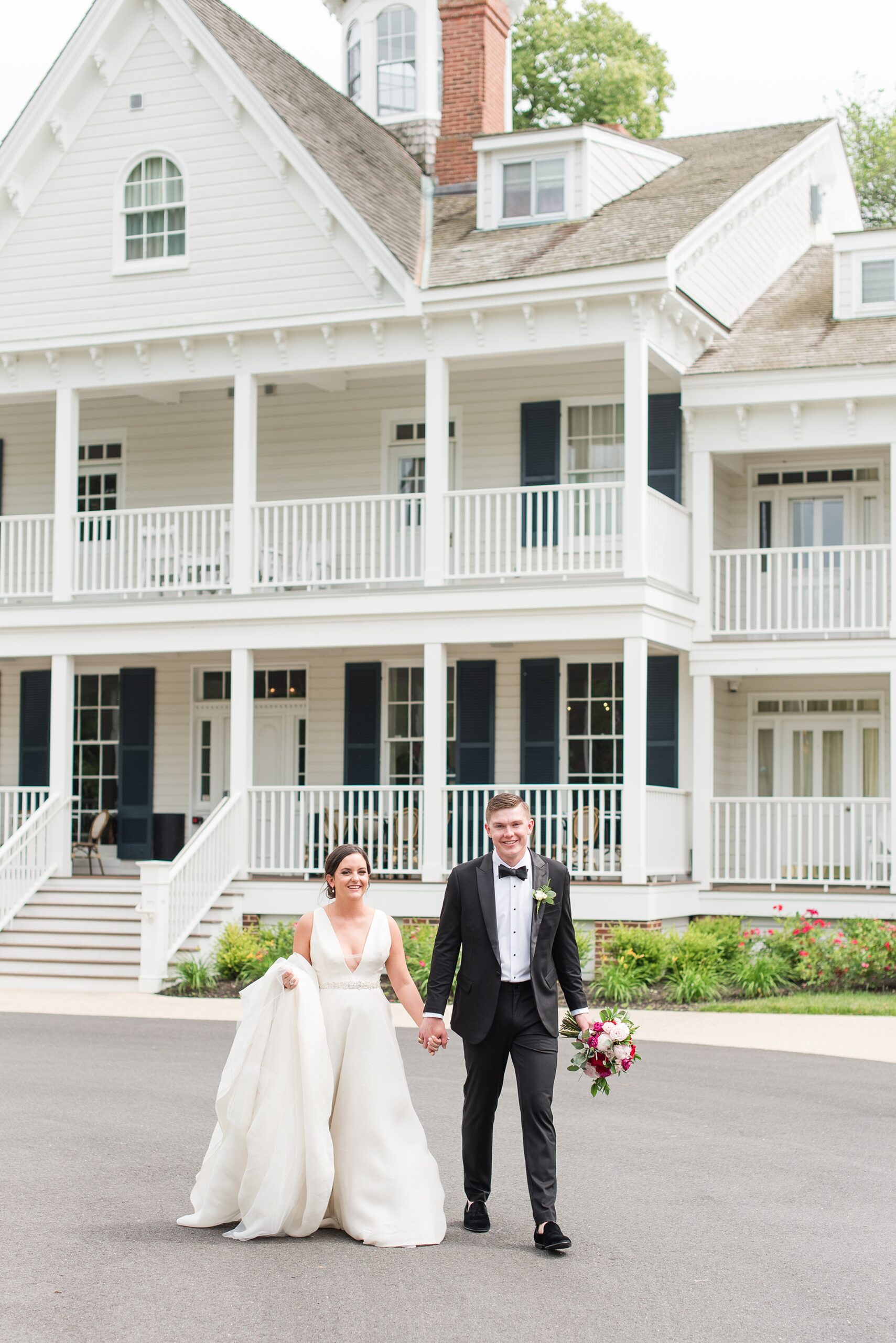 Newlyweds hold hands while walking through the driveway in front of a white building