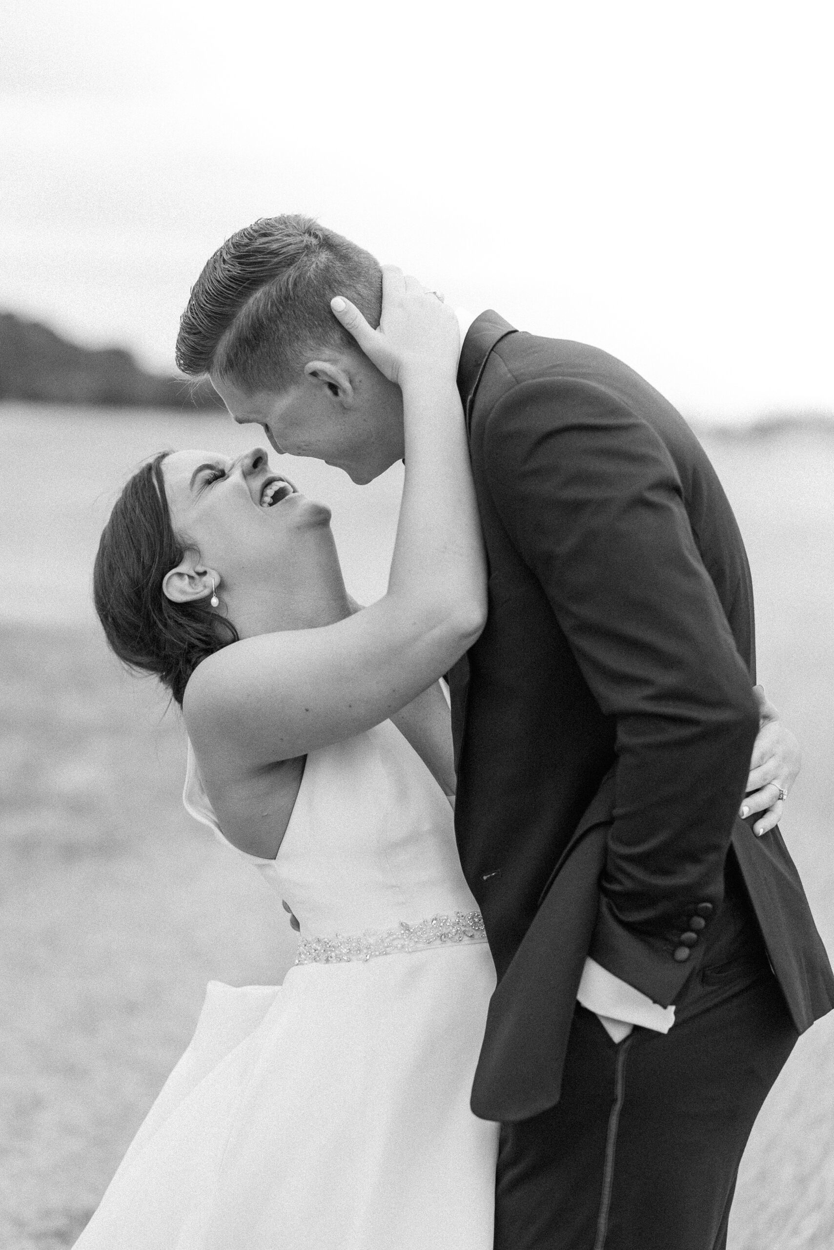 A bride laughs while her groom leans in for a kiss in an open field