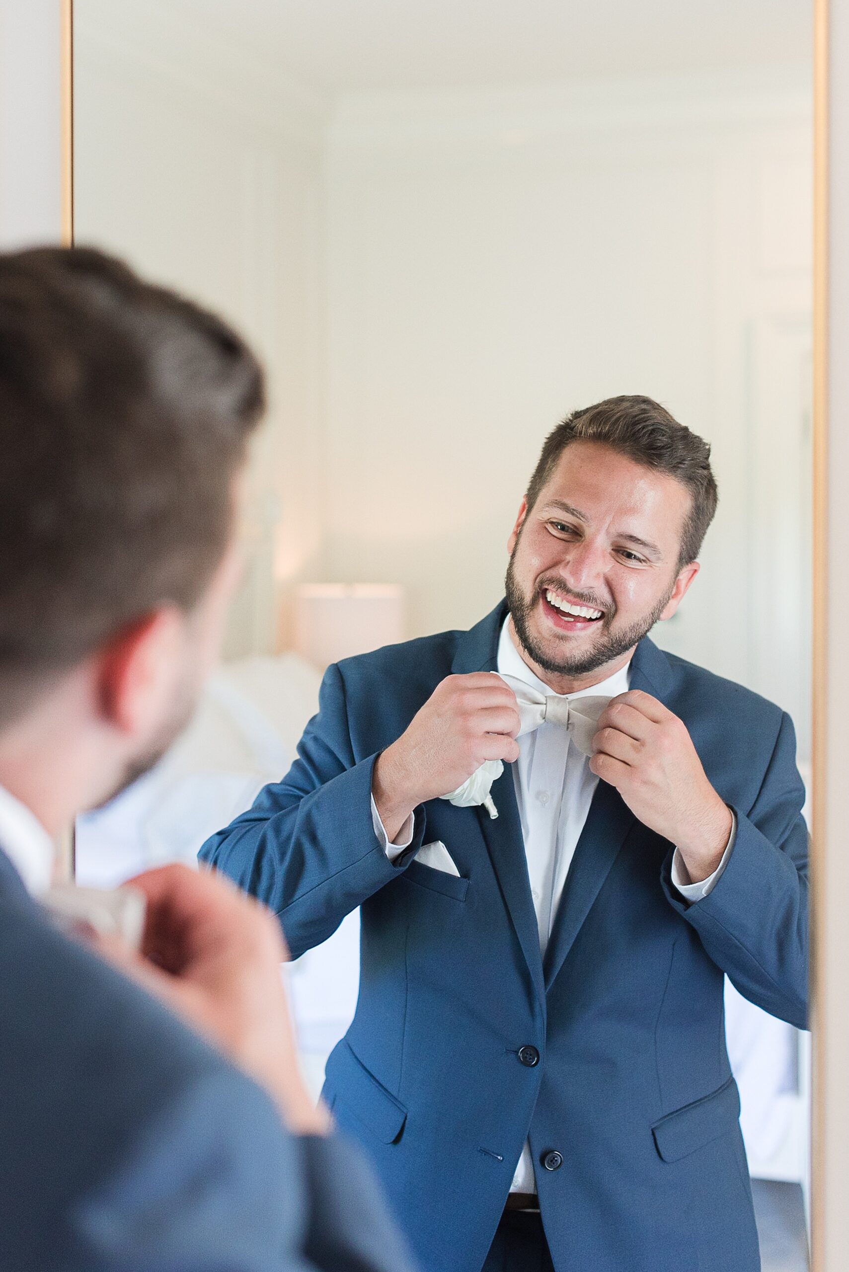A groom laughs while adjusting his white bowtie in a mirror