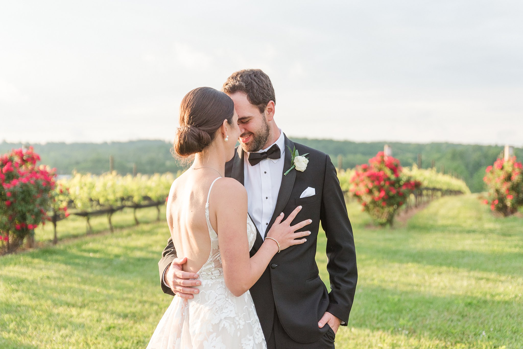 Newlyweds stand in a vineyard touching foreheads and smiling at sunset