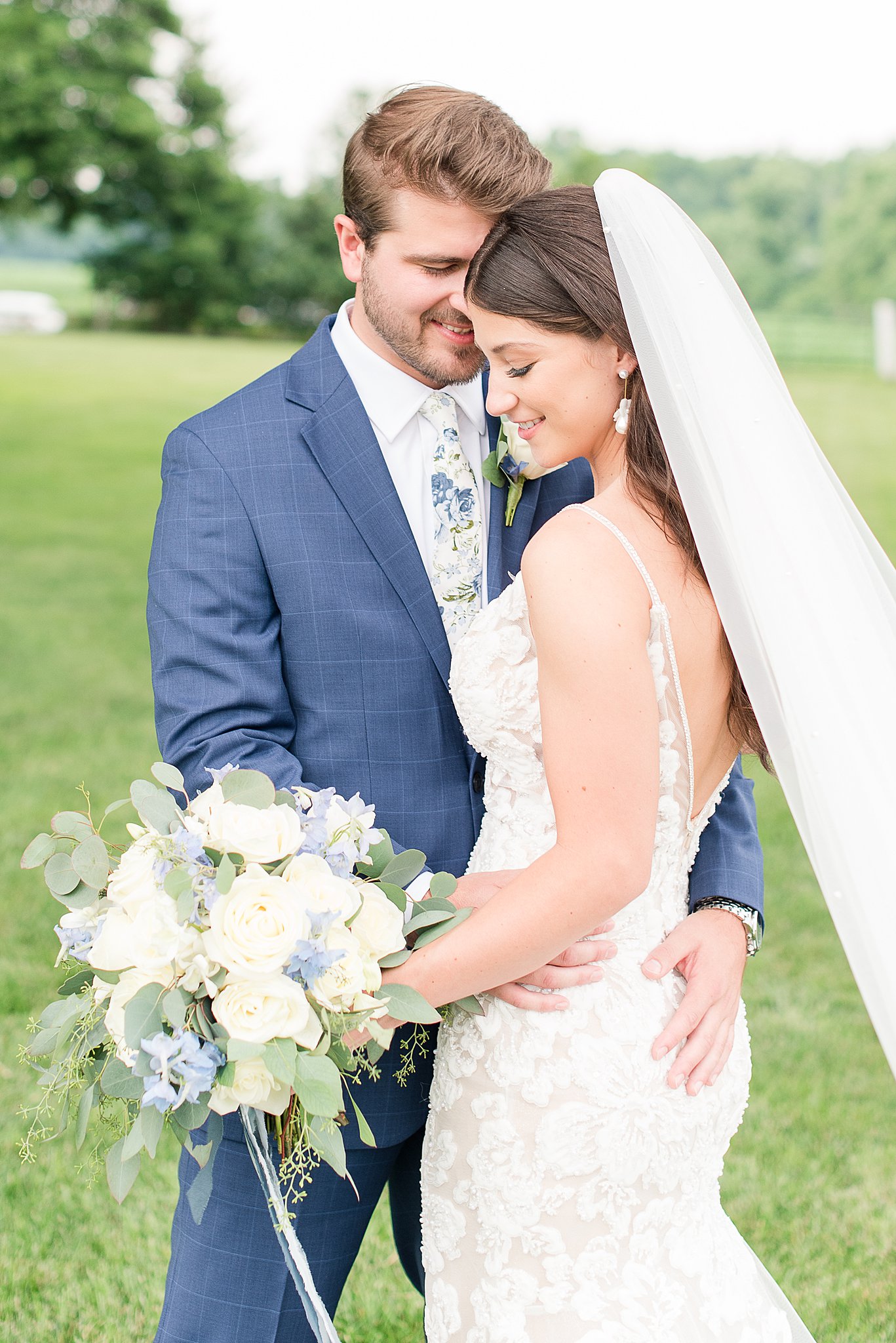 Newlyweds stand close while wearing a lace embroidered dress and blue plaid suit in a lawn at one of the Outdoor Wedding Venues In Baltimore