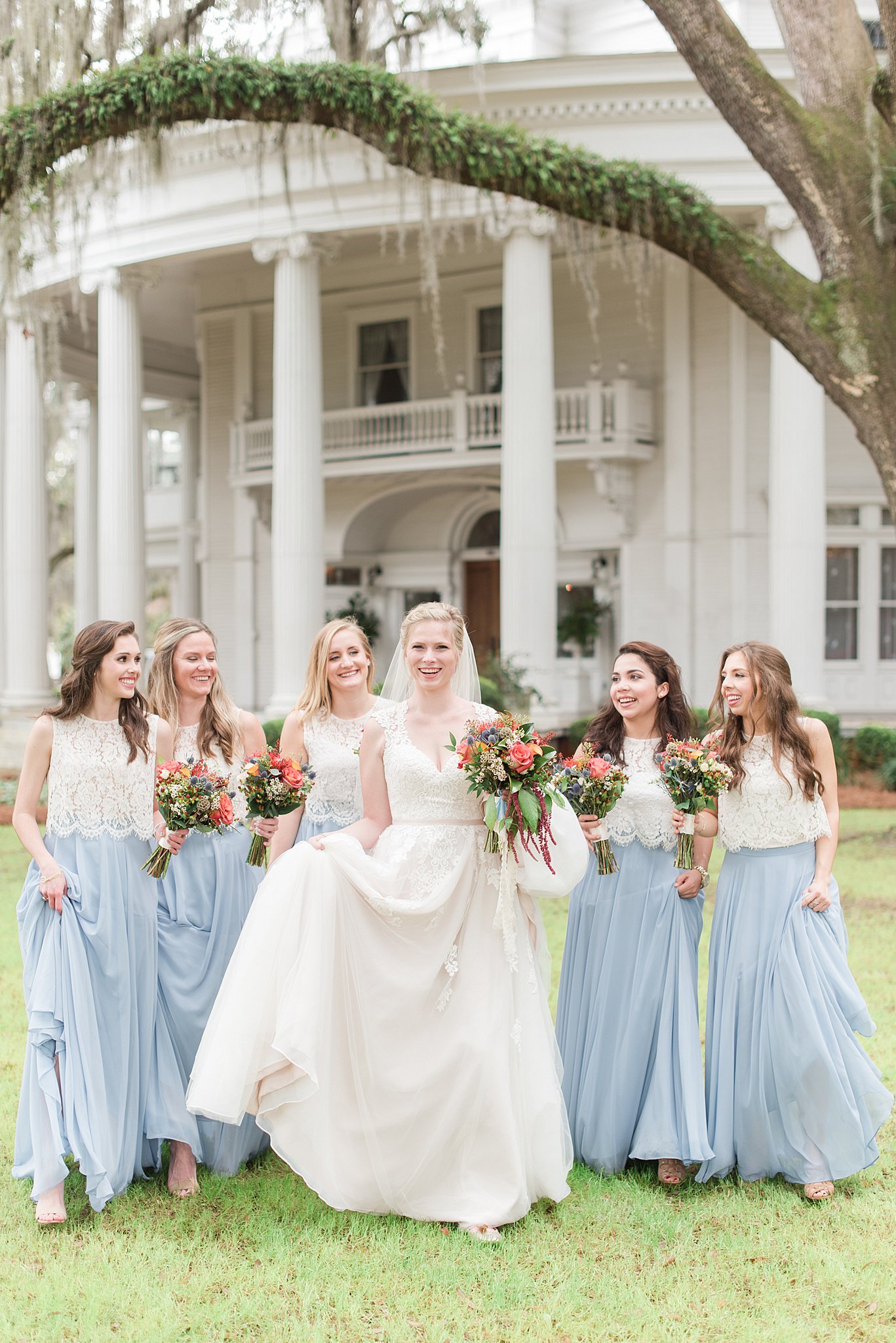 A bride walks through a lawn under oak trees while surrounded by her bridal party in blue and white dresses