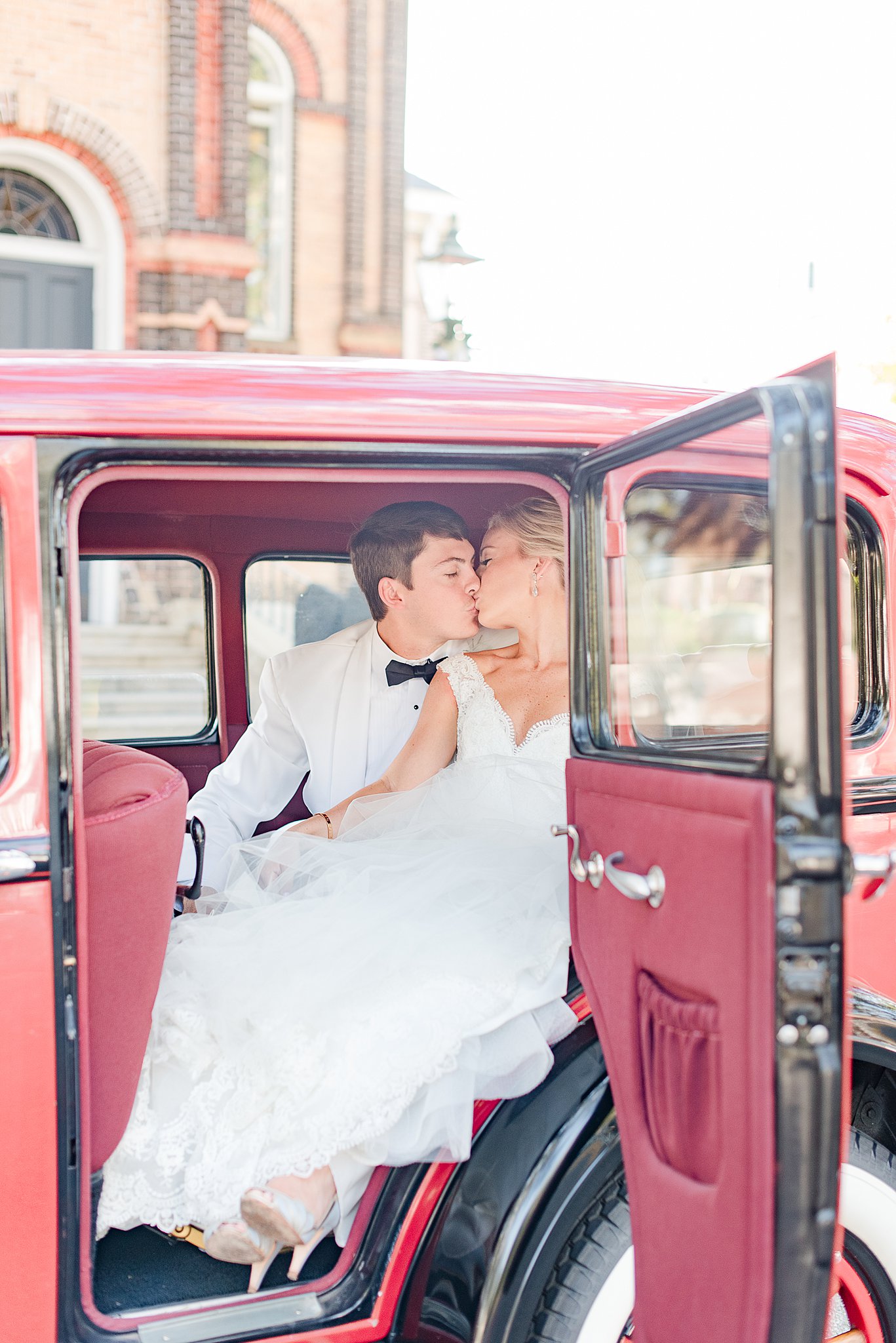 Newlyweds in white suit and lace dress kiss while sitting in the back of a vintage red car