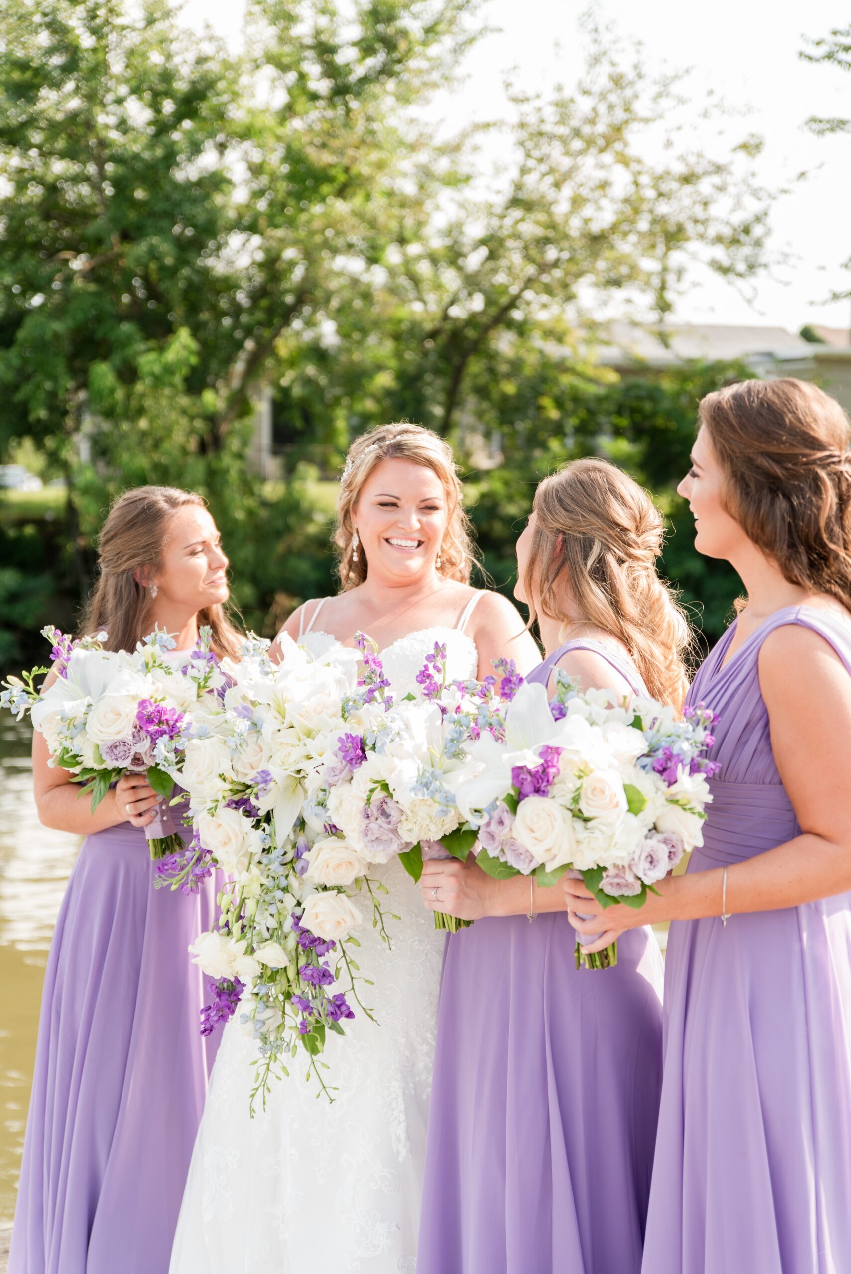 A bride laughs while standing on a dock with her bridesmaids in purple dresses