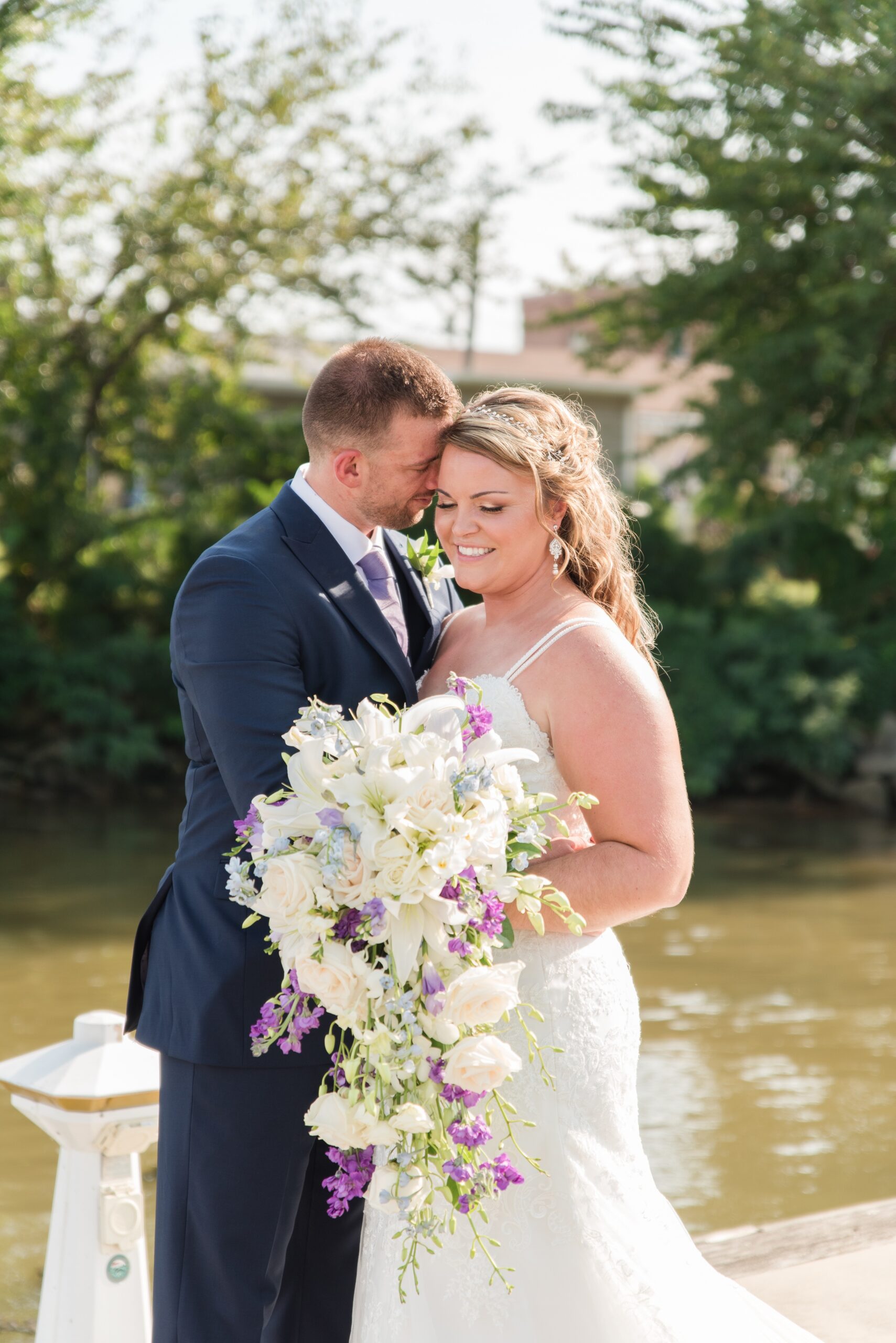 A groom nuzzles his bride while they stand on a dock over the water