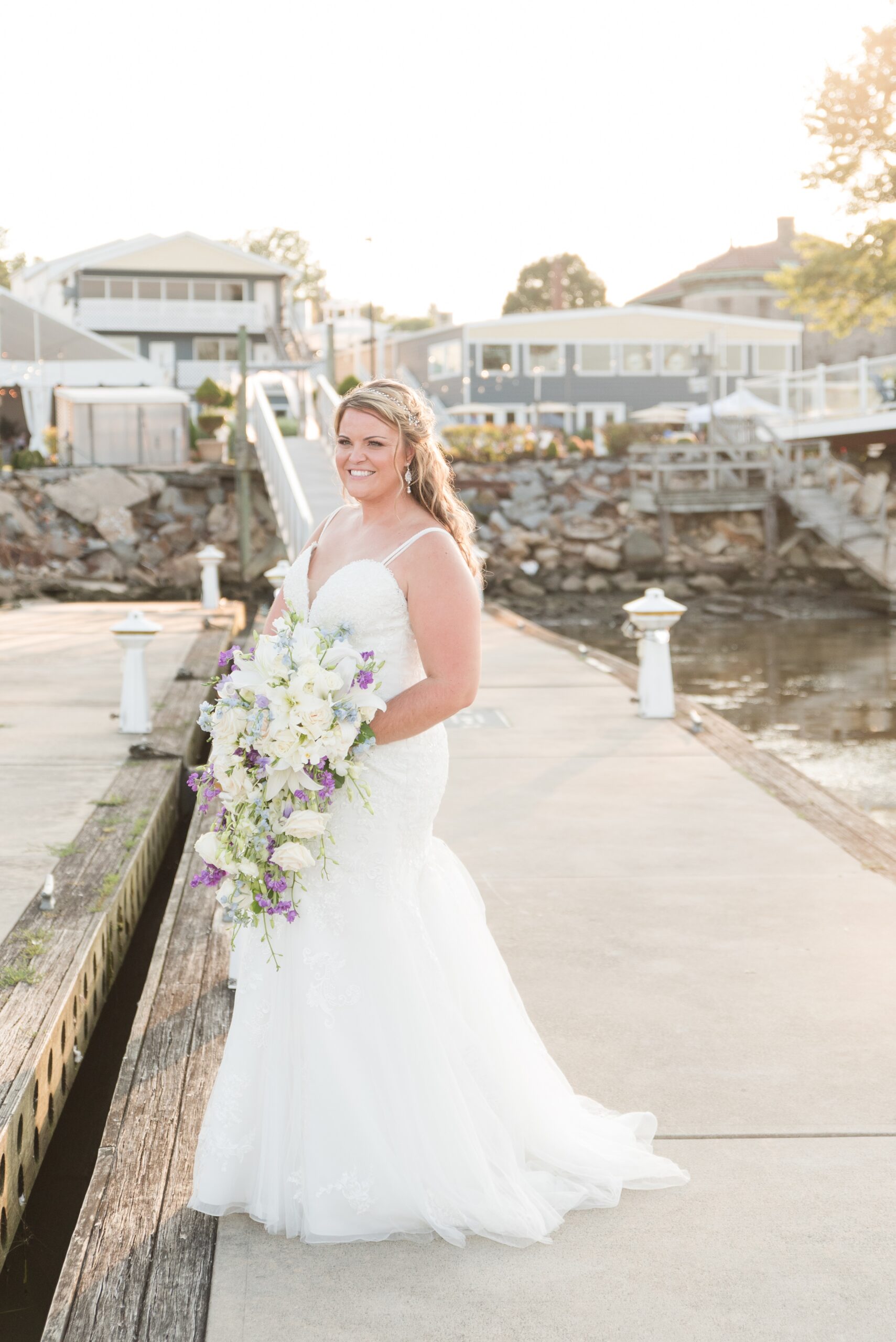 A bride smiles while standing on a dock and holding her large falling white and purple bouquet at her La Banque de Fleuve Wedding