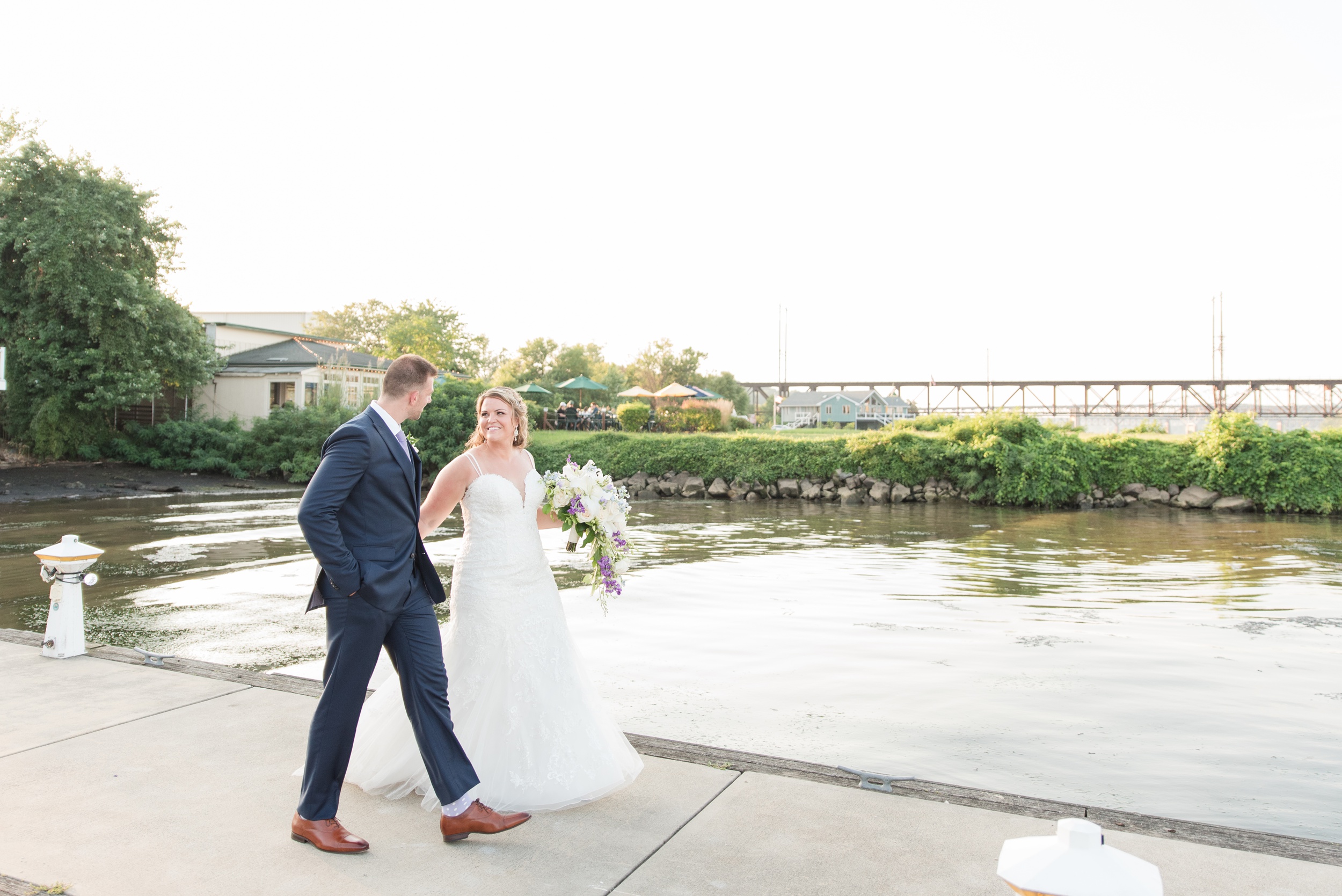 A bride and groom walk up a long dock over the water at their La Banque de Fleuve Wedding
