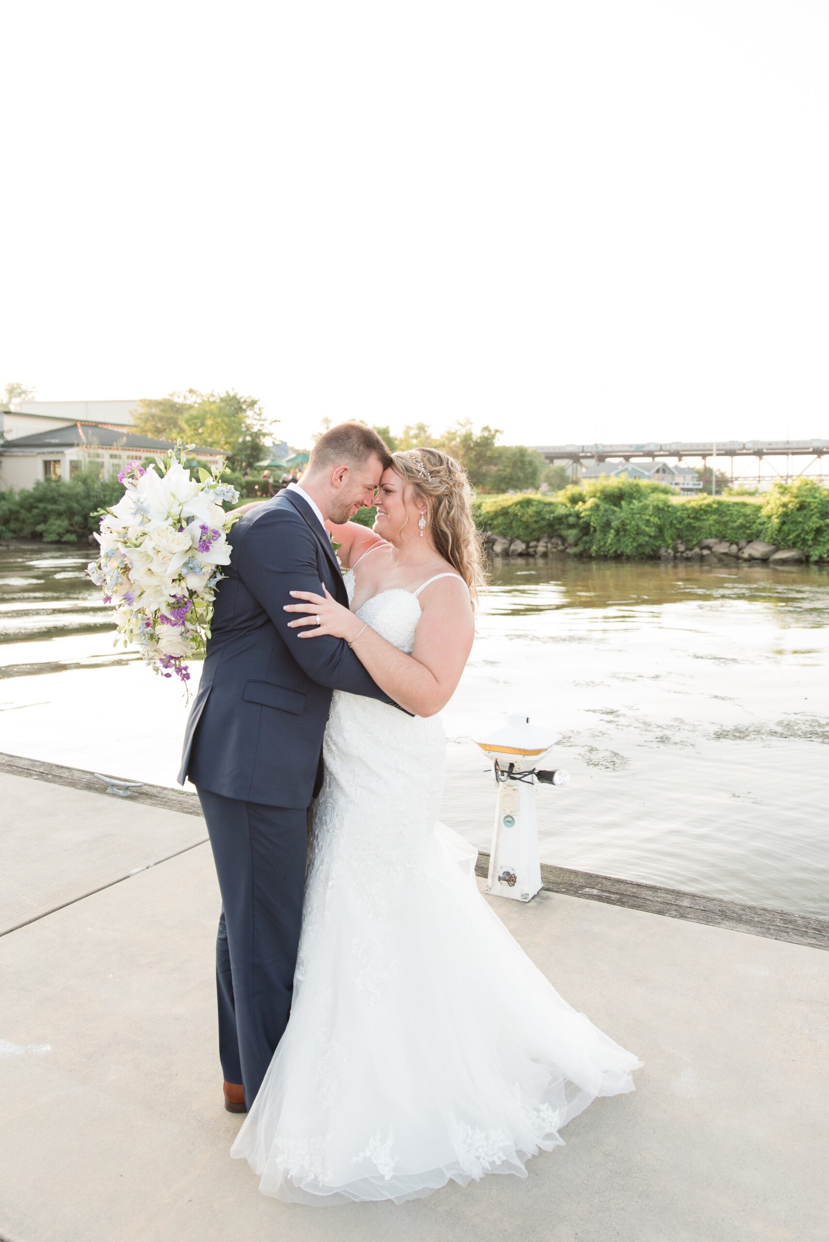 Newlyweds touch foreheads while standing on a dock and holding a large purple and white bouquet