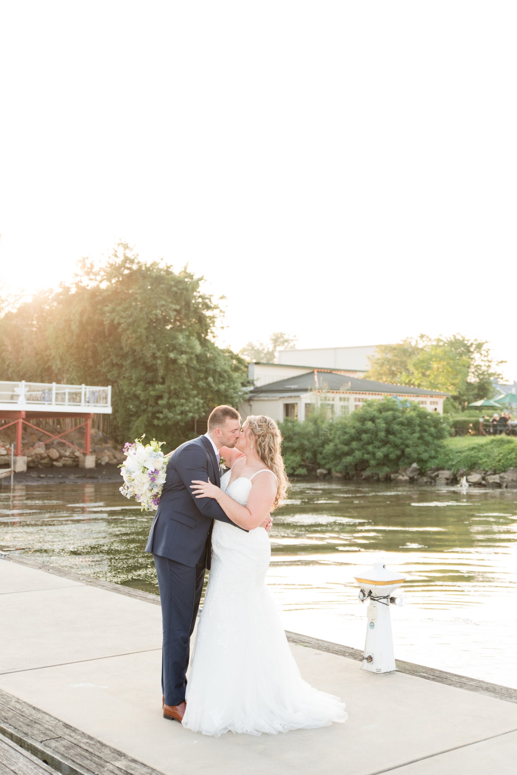 A bride and groom kiss while standing on a dock at sunset during their La Banque de Fleuve Wedding