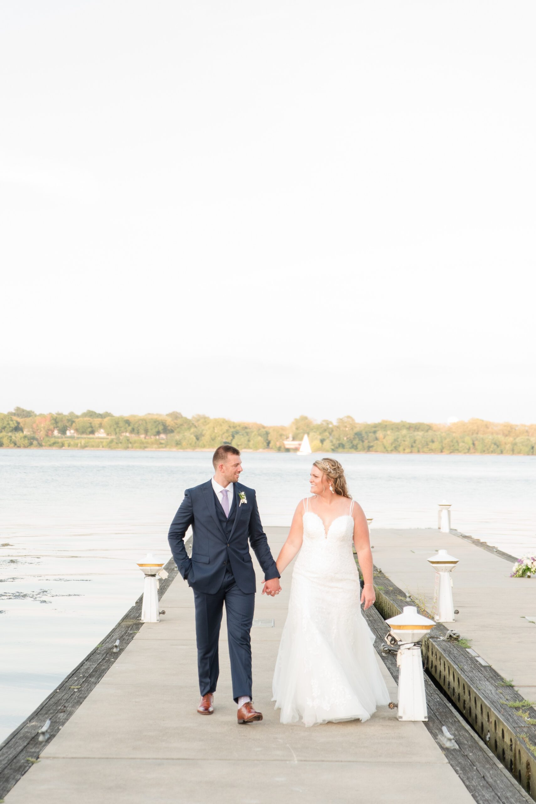 A bride and groom walk up a dock holding hands and smiling at each other at sunset during their La Banque de Fleuve Wedding