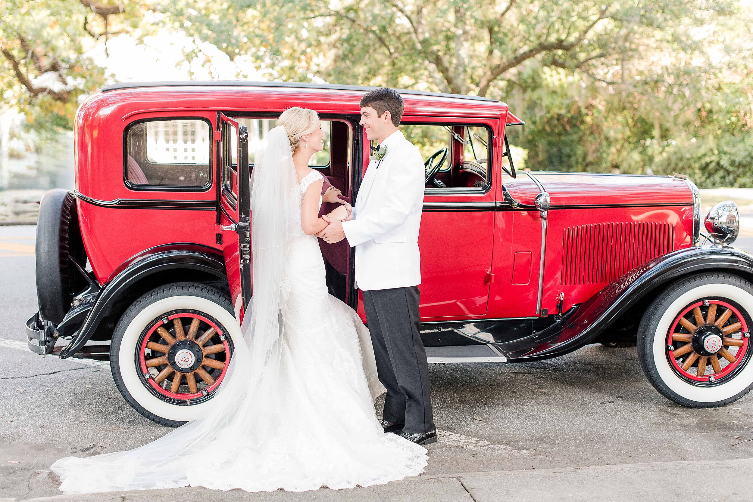 A groom in a white suit helps his bride into a red vintage car at their Manor Country Club Wedding