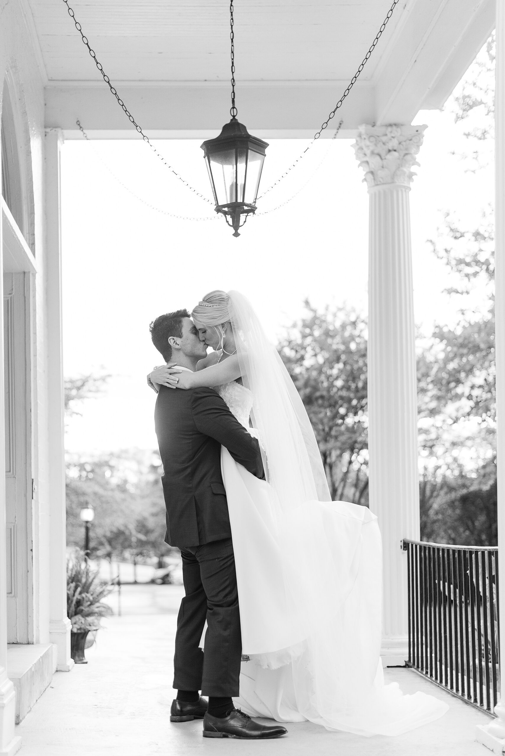 A groom lifts and kisses his bride on a white porch under a hanging light at their Sagamore Pendry Baltimore wedding