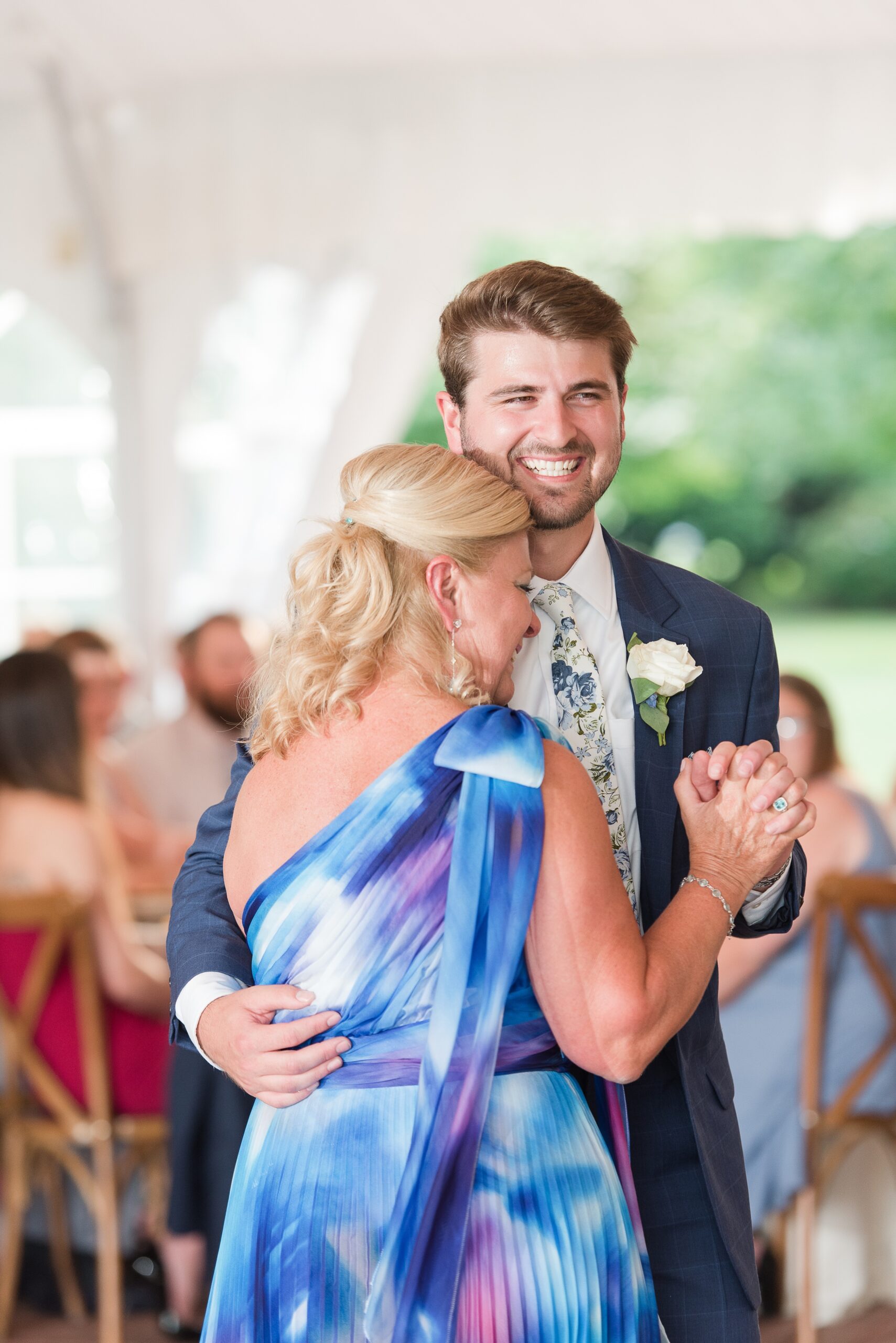 A groom in a blue suit dances with him mom with big smiles during his wedding reception