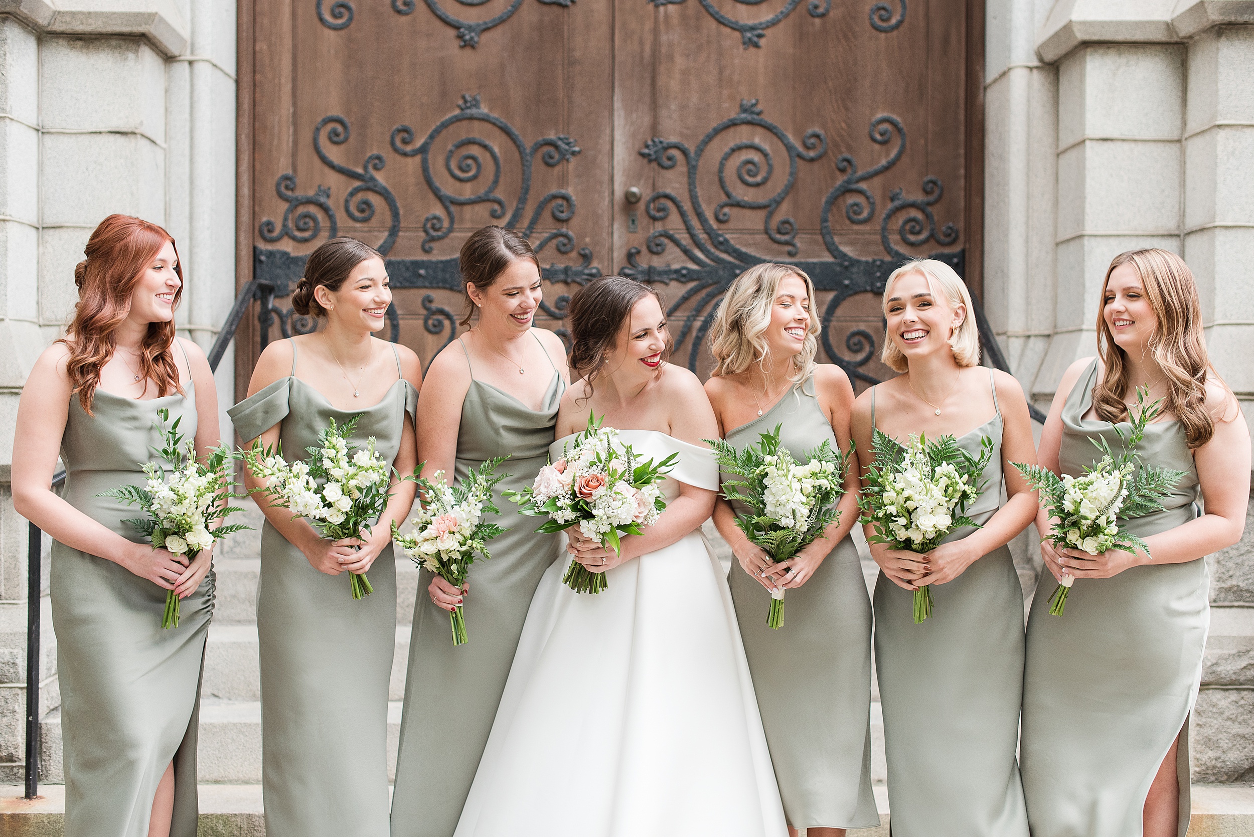 A bride laughs with her bridesmaids while standing on the front steps of an ornate wooden door entrance