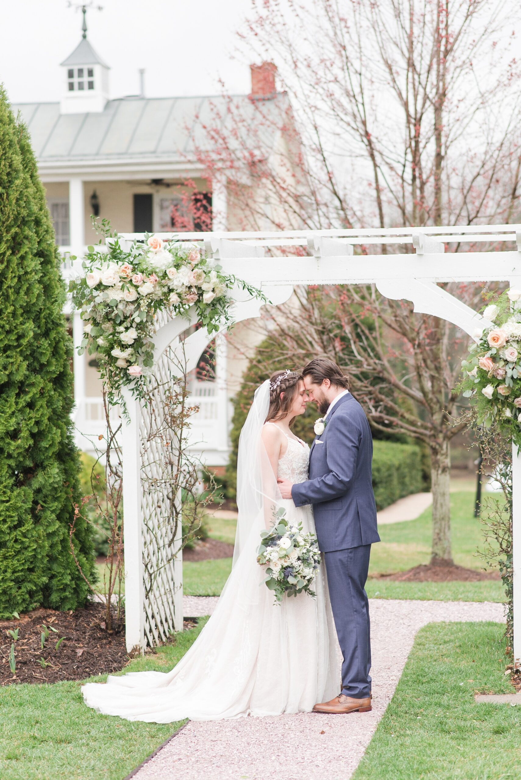 Newlyweds touch foreheads while standing under a white pergola