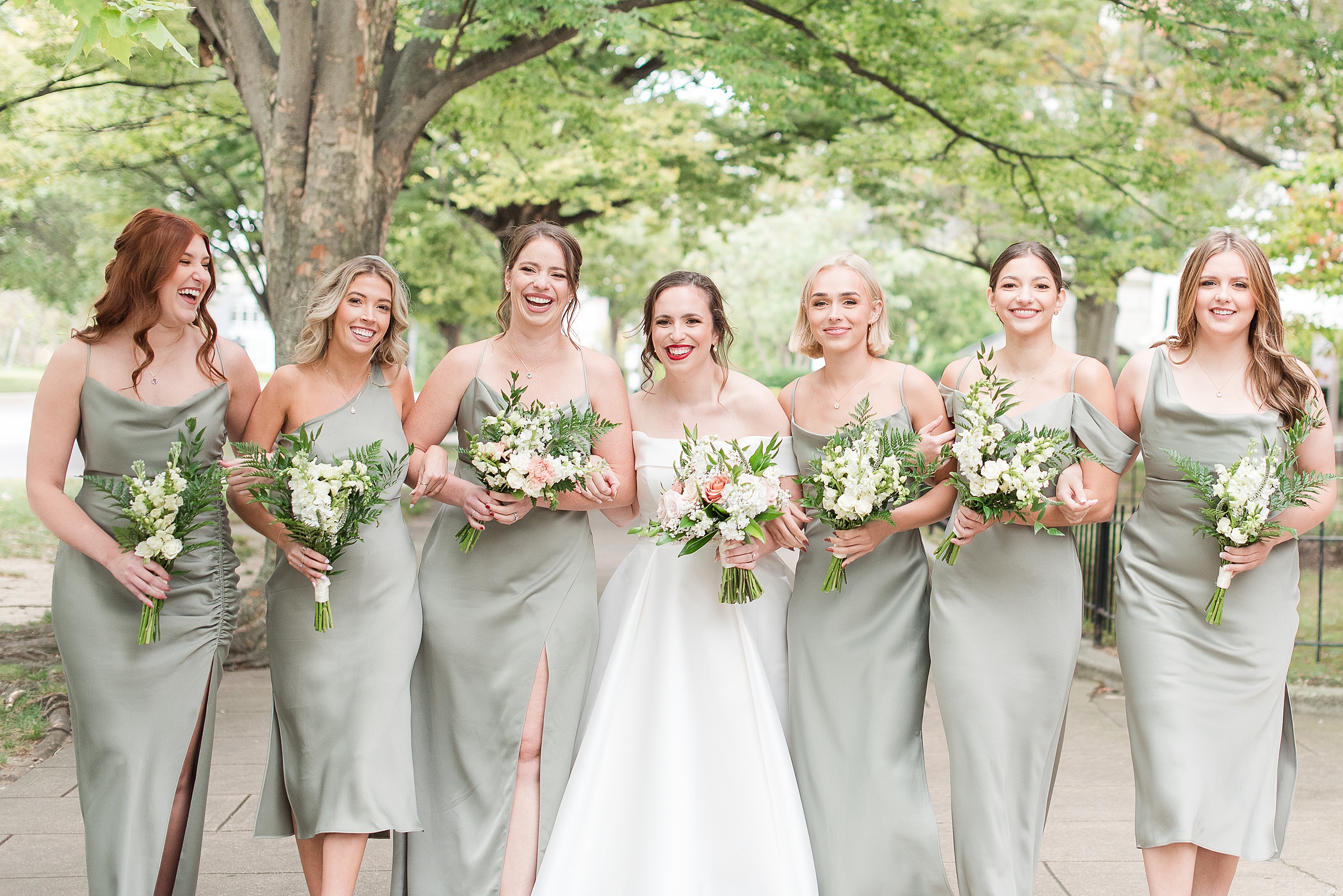 A bride laughs with her bridesmaids in green dresses while walking up a sidewalk