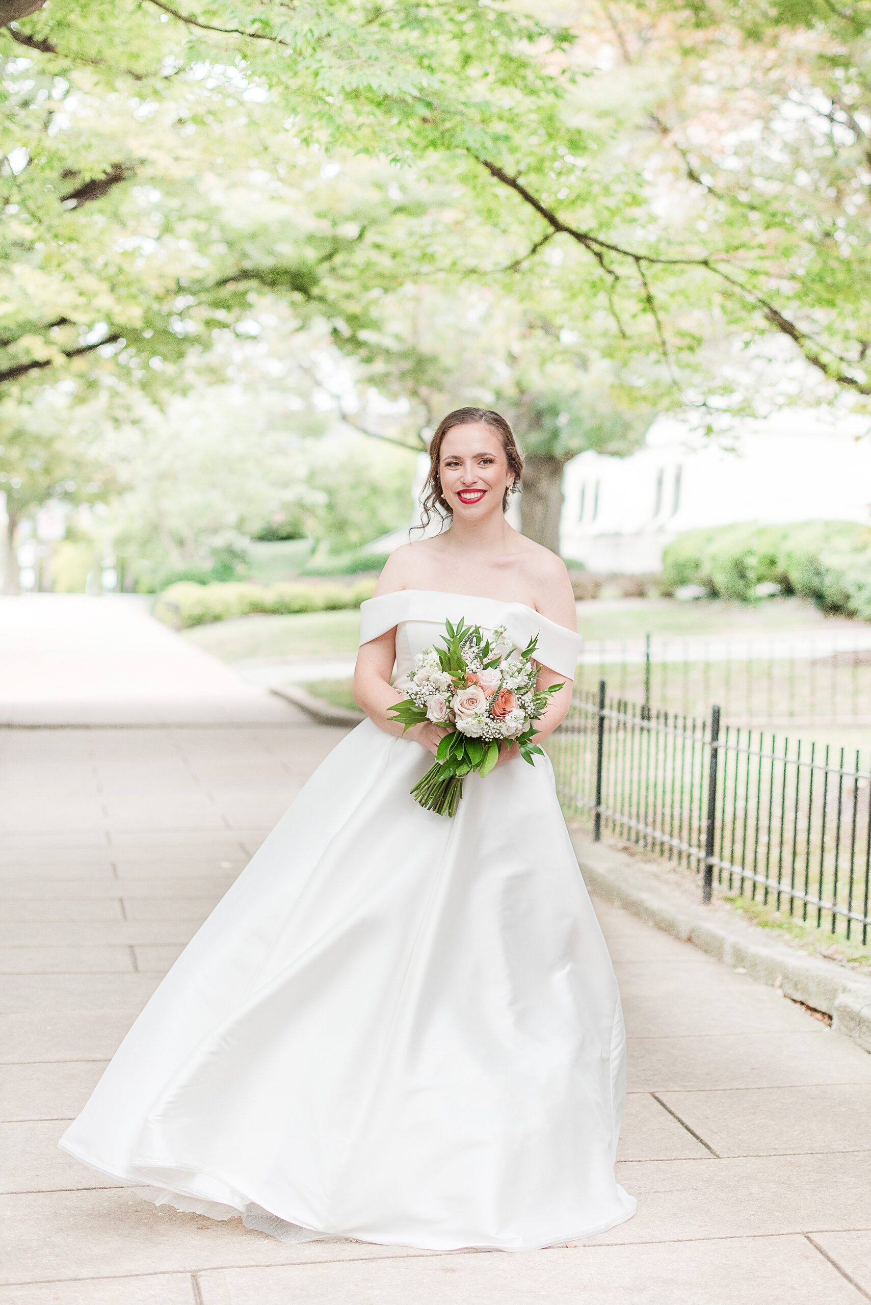 A bride in a silk dress stands on a windy sidewalk holding her bouquet and smiling at her Baltimore Country Club Wedding