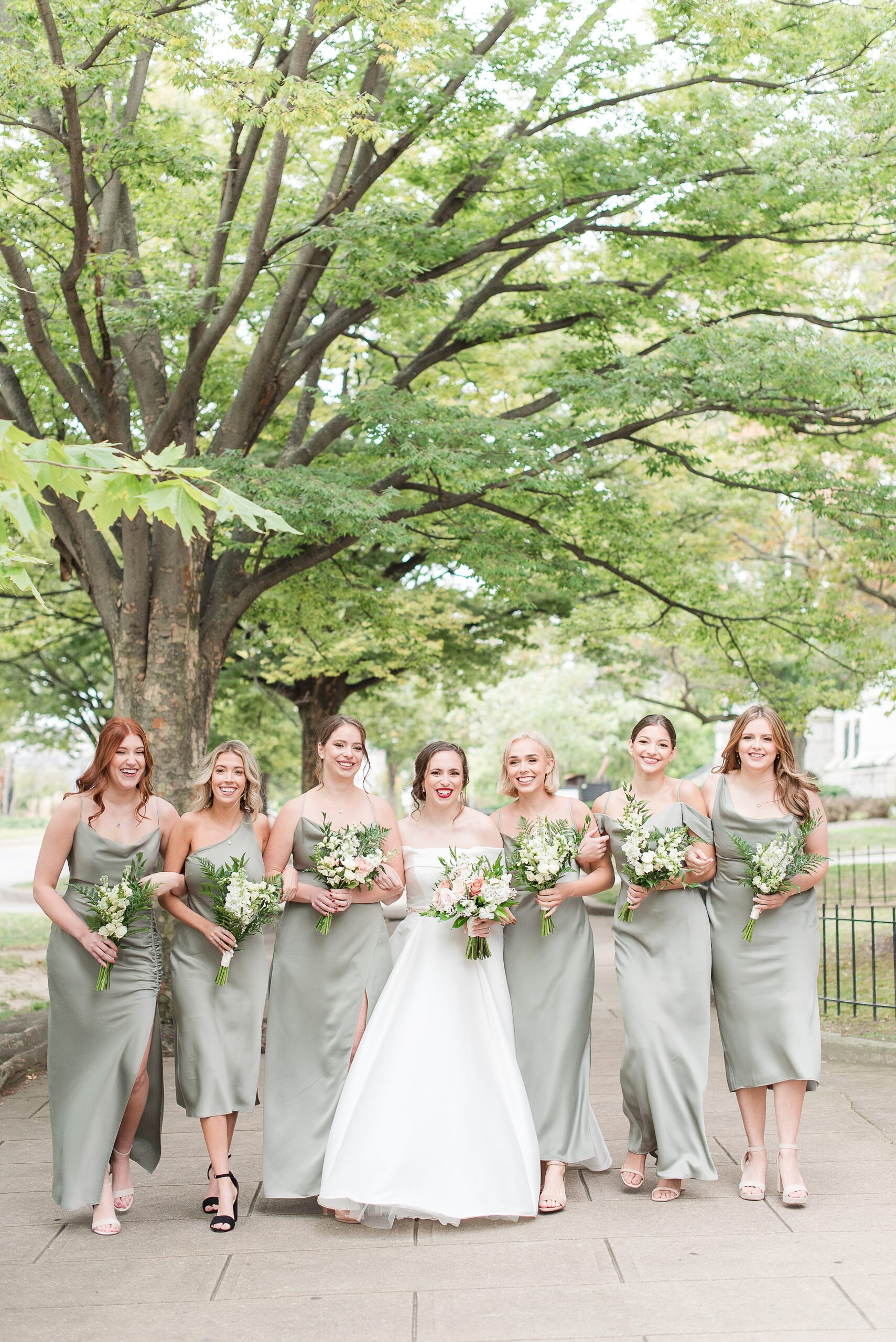 A bride walks down a manicured sidewalk with arms locked under trees at her Baltimore Country Club Wedding