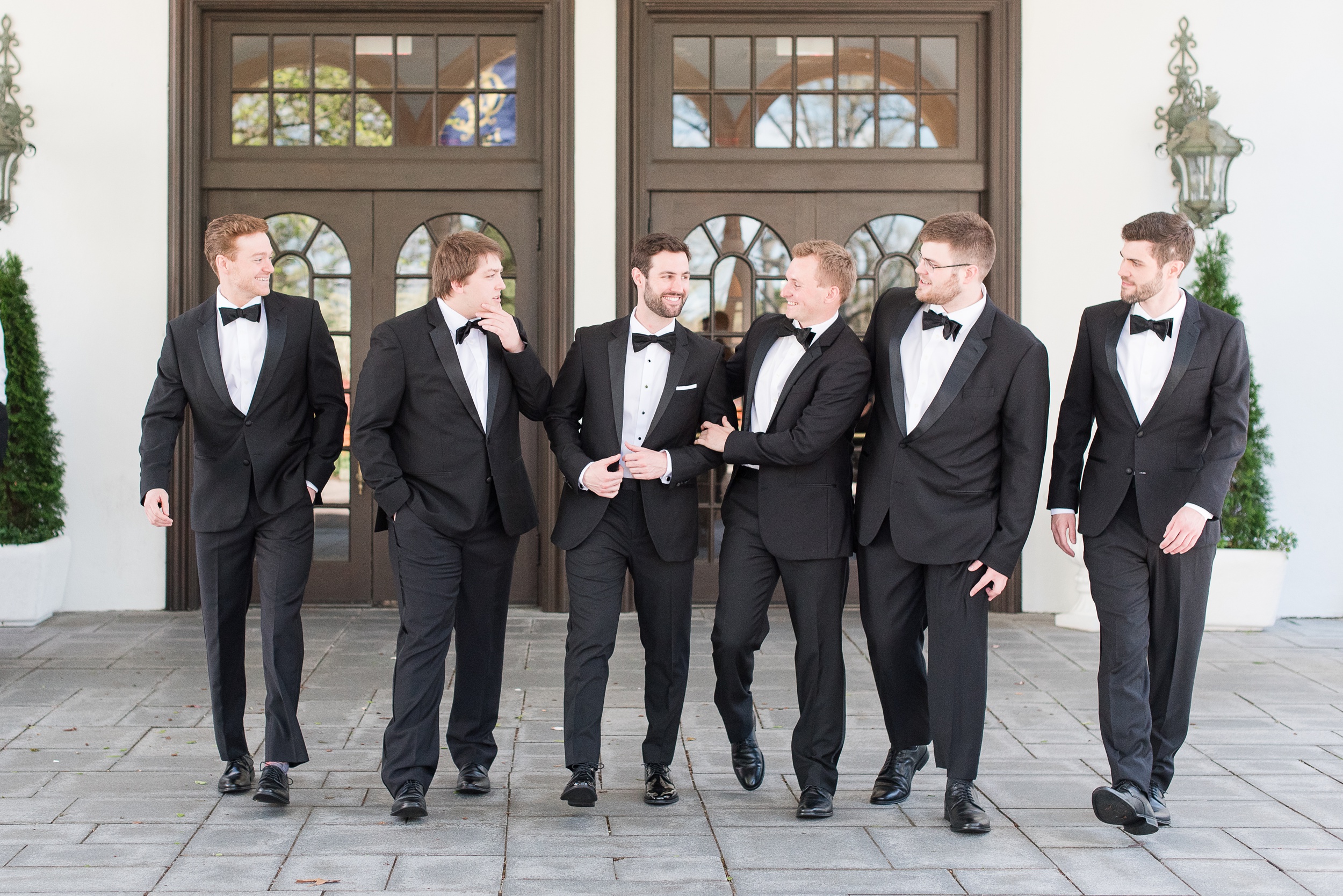 A groom walks on a patio while laughing with his groomsmen in matching black tuxedos