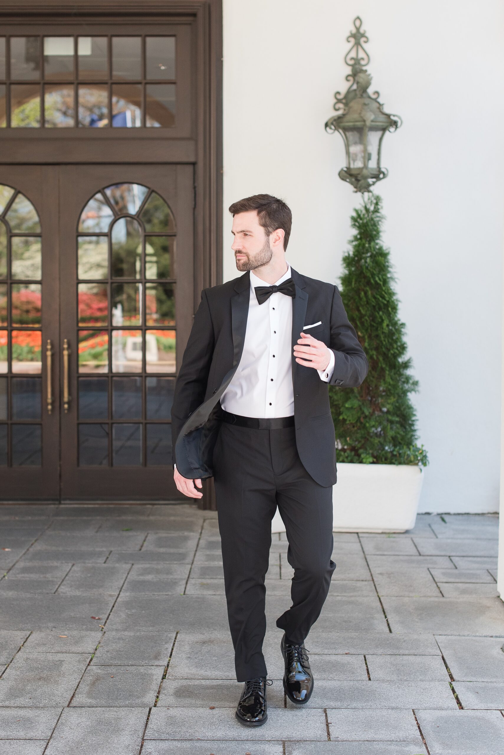 A groom in a black tuxedo walks on a stone patio with a hand on his lapel