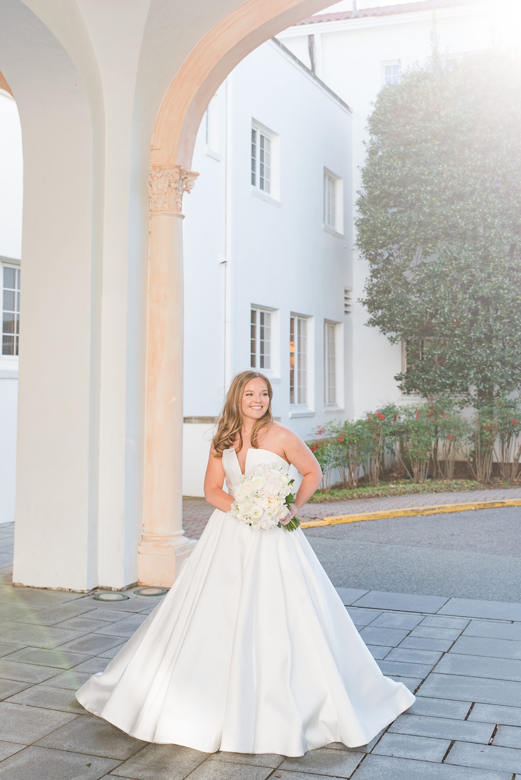 A bride smiles over her shoulder while standing in a car port holding her white bouquet
