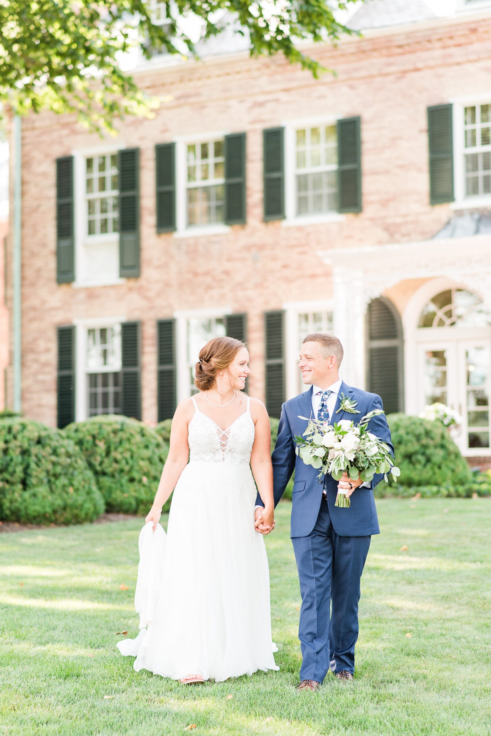 A bride and groom walk hand in hand through the lawn while the groom holds the bouquet