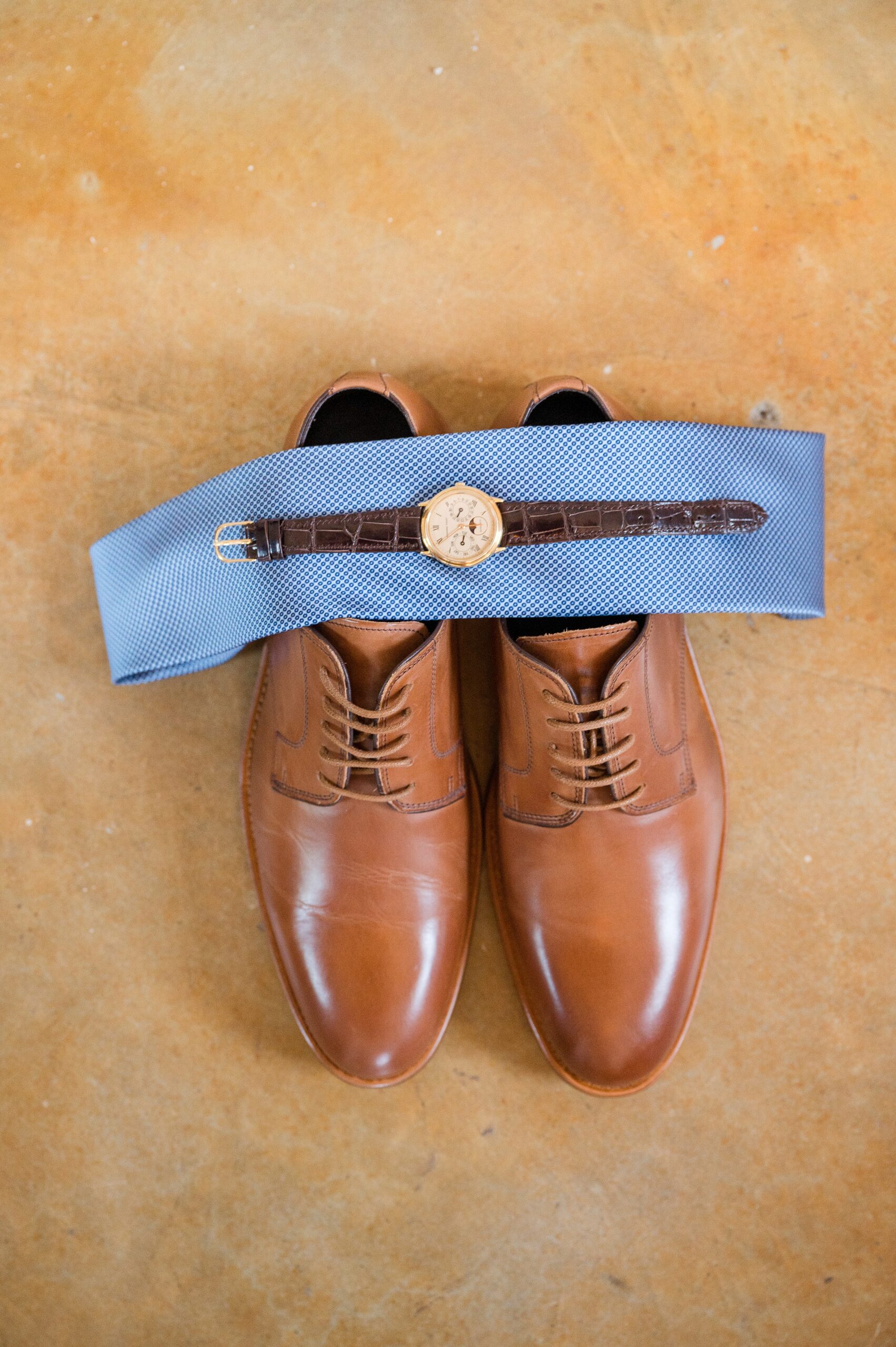 Groom details of shoes, tie and watch on a brown floor