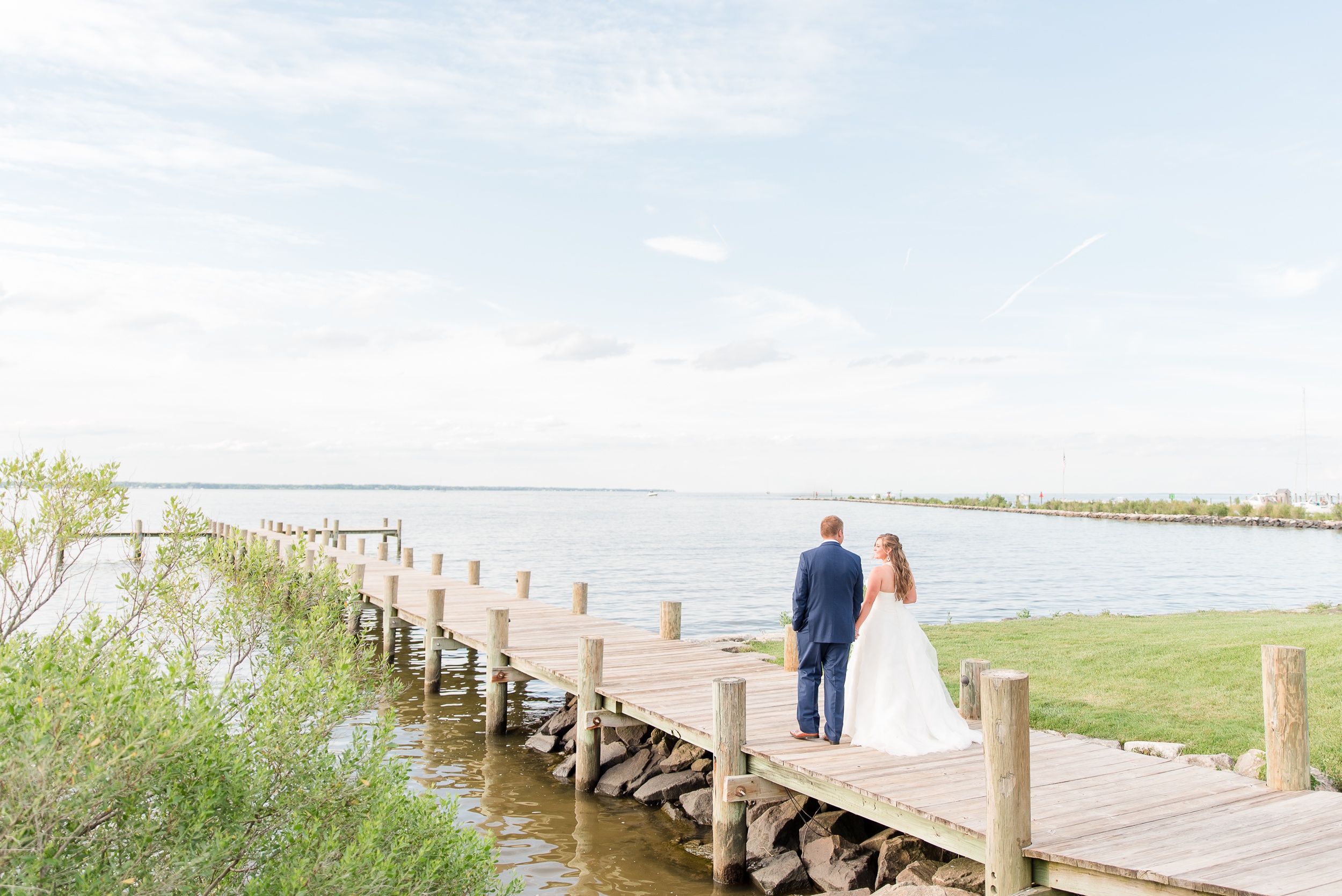 Newlyweds smile at each other while holding hands and walking on a long wooden dock during their Herrington On The Bay Wedding