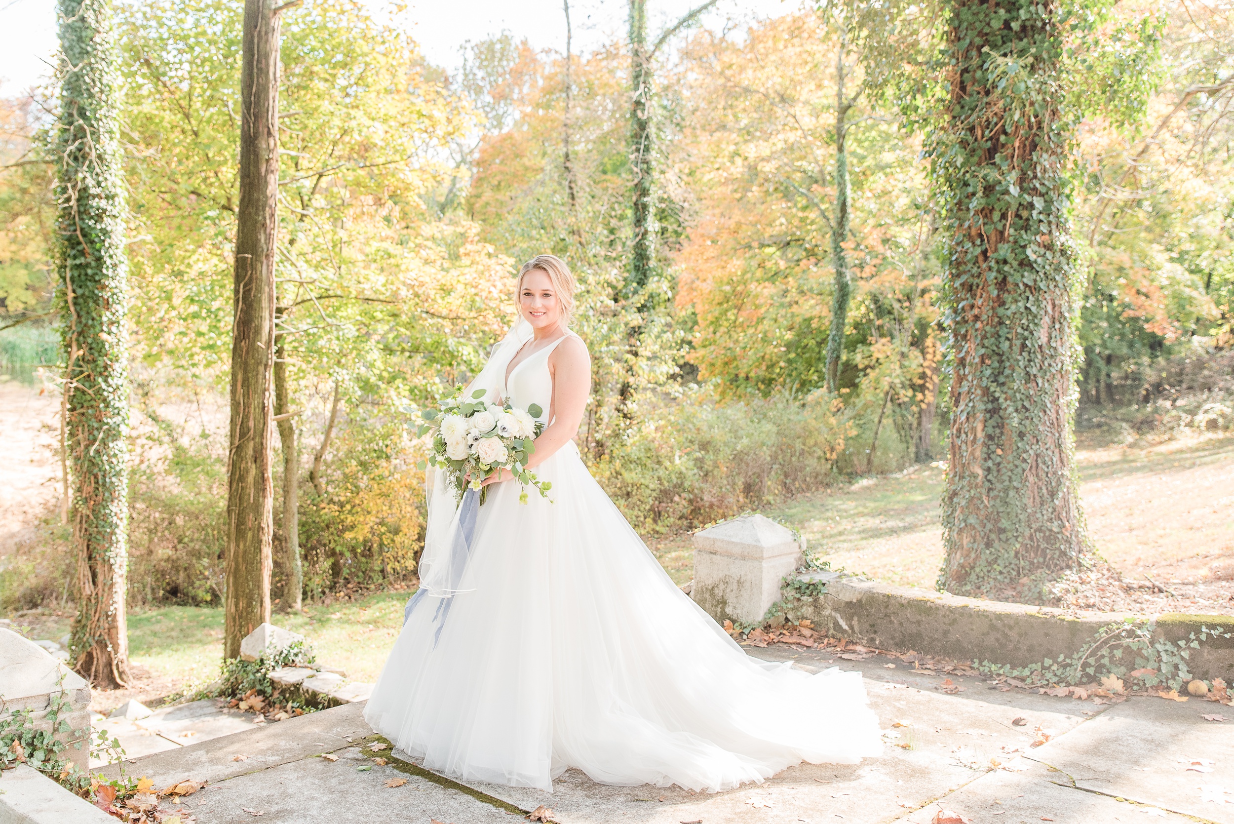 A bride stands in a garden path holding a white rose bouquet and smiling at her Historic Shady Lane Wedding