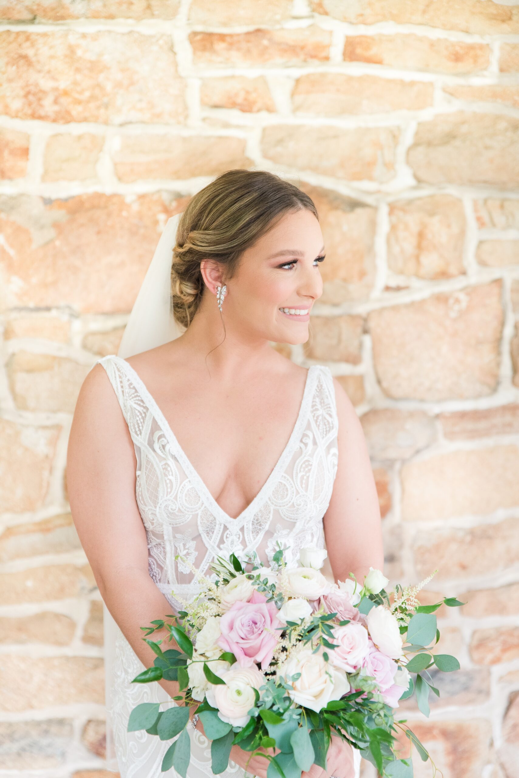 A bride leans on a stone wall in a lace dress while smiling and holding her pink and white rose bouquet